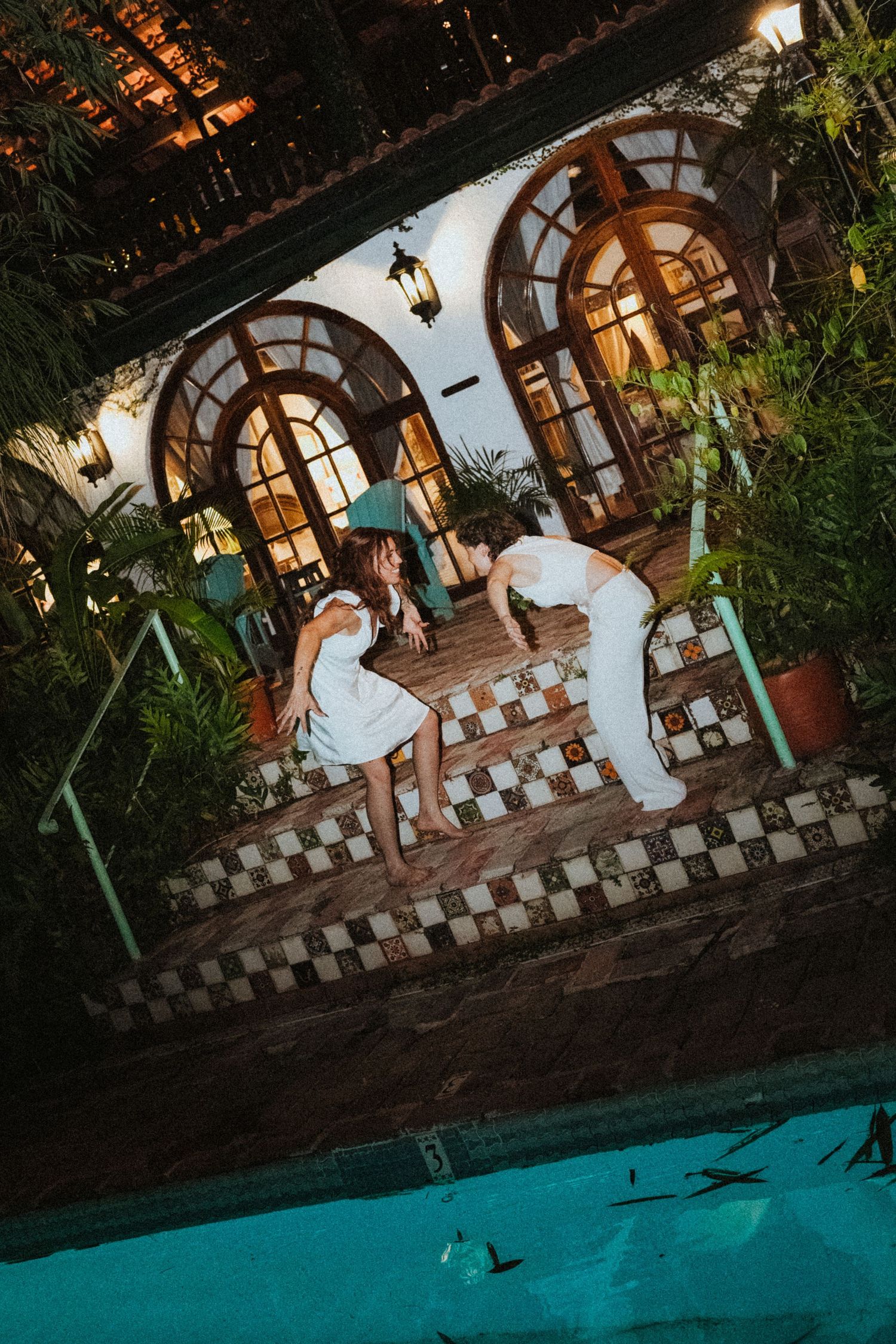 Two women in white dresses pose on tiled steps beside a pool at an elegant tropical venue at night.