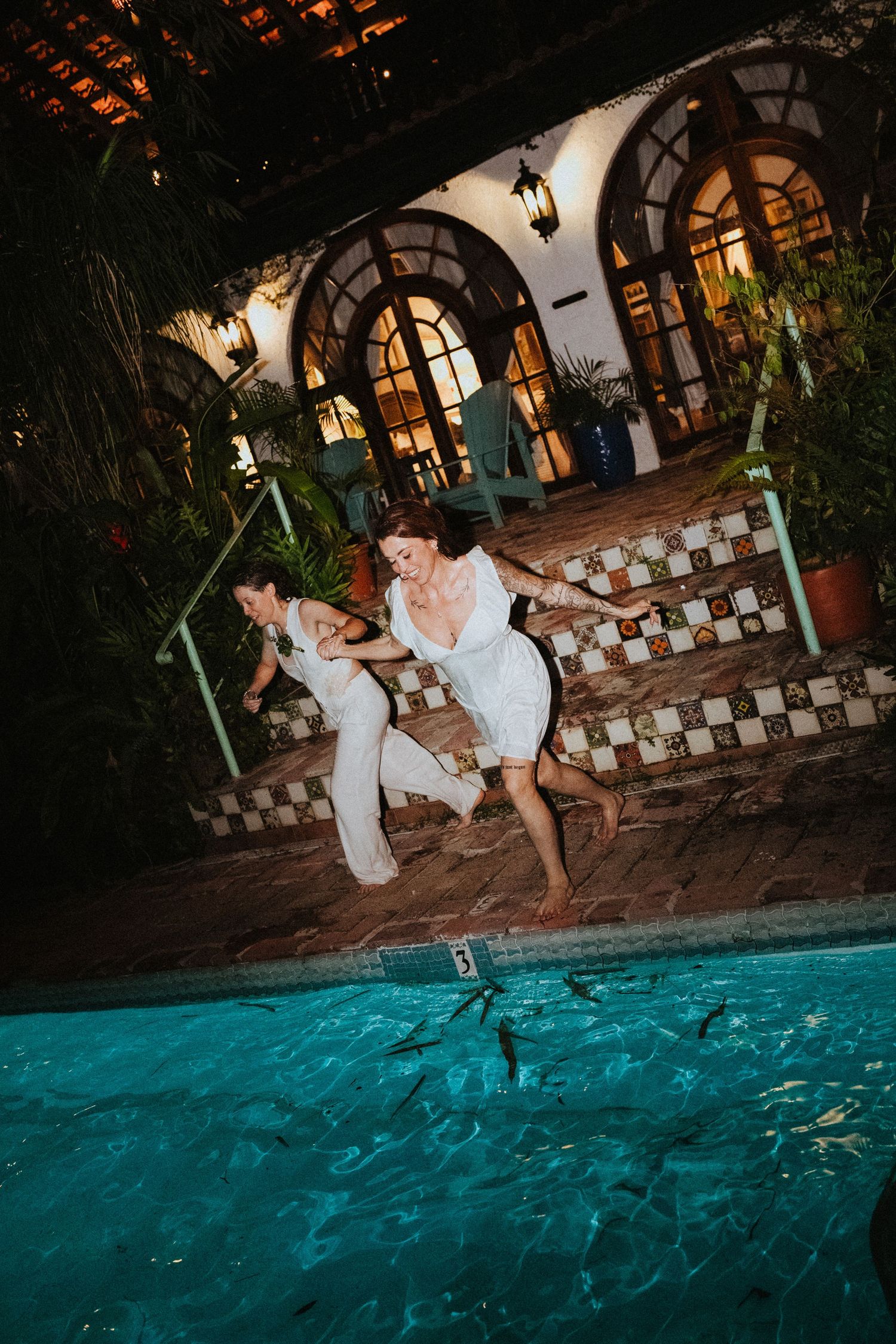 Two brides in white dresses joyfully jumping into a pool at night at an elegant venue.