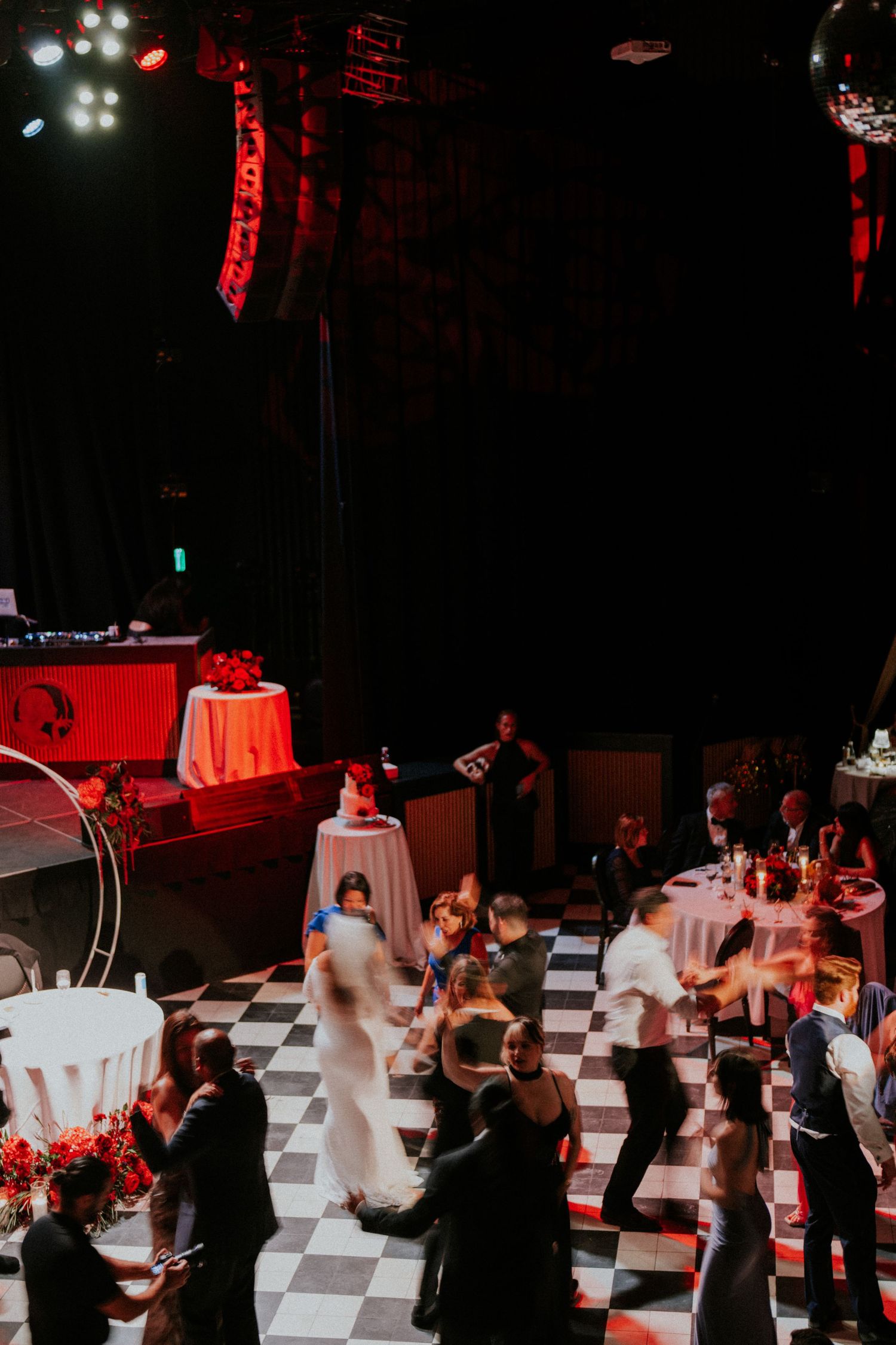 Guests dancing on a black and white checkered dance floor at an elegant wedding reception with red lighting.