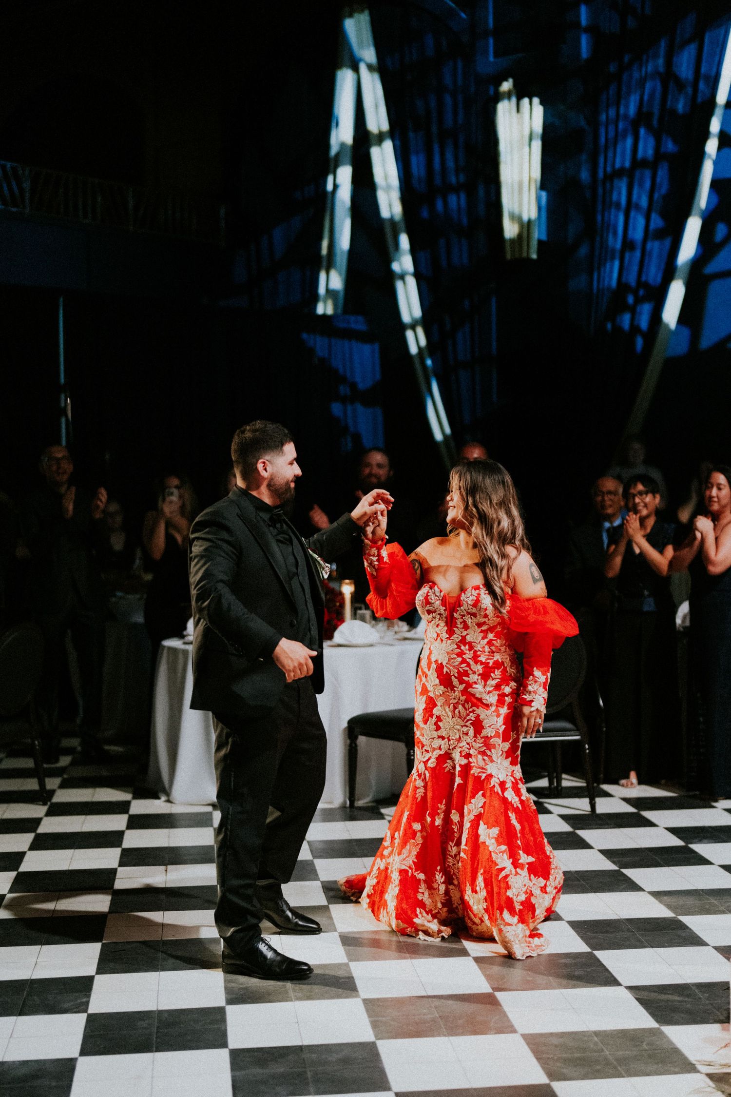 Couple shares first dance on checkered floor, bride in red lace mermaid gown, groom in black suit.