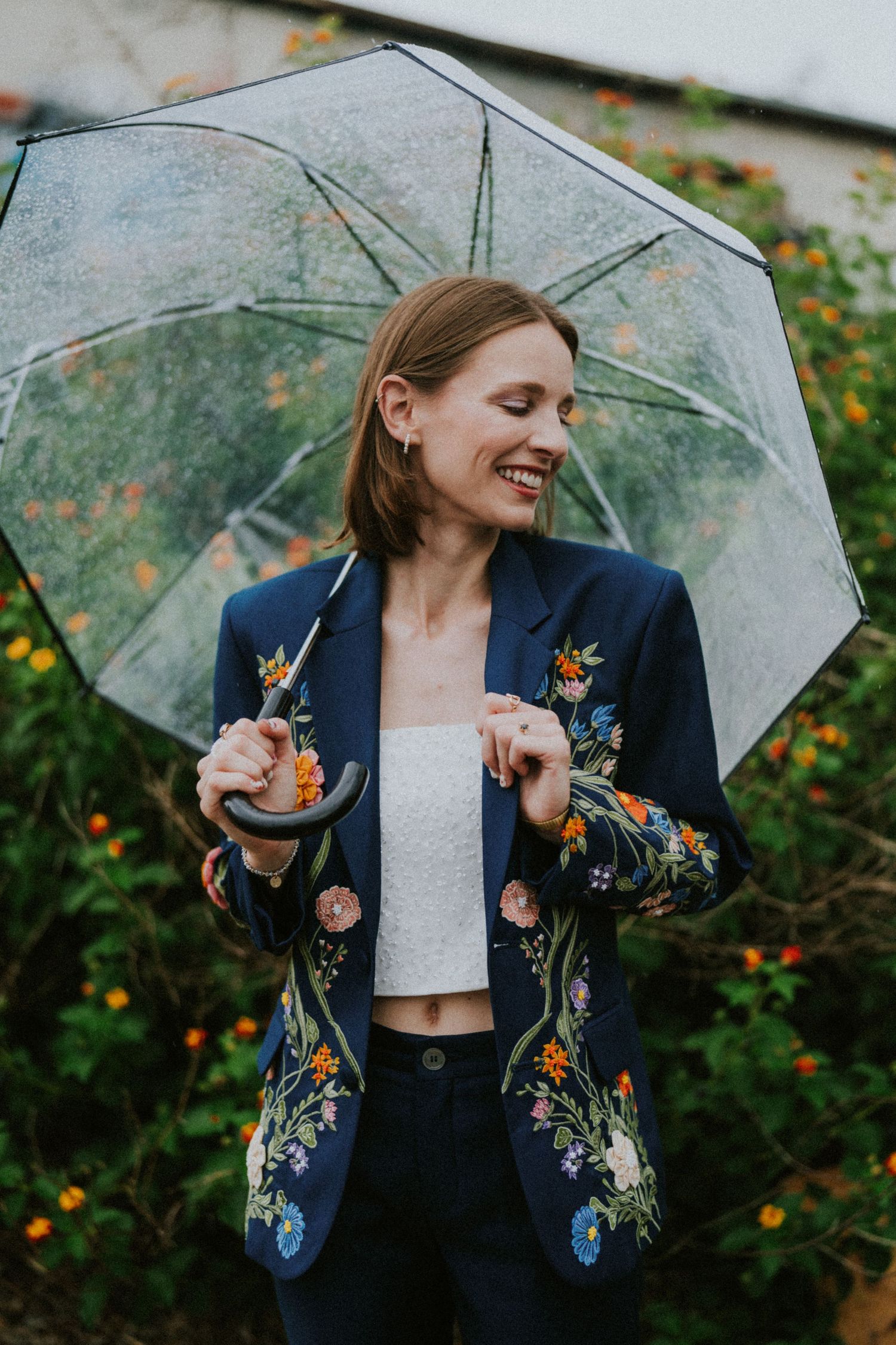 Smiling woman in floral embroidered navy blazer holding a clear umbrella outdoors among colorful flowers.