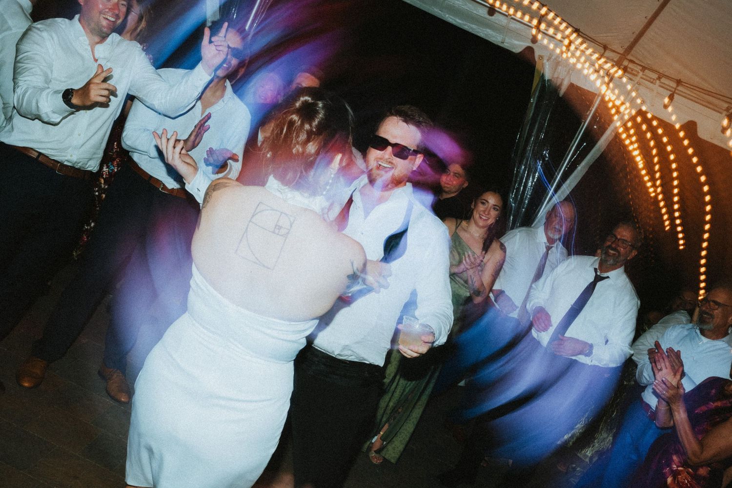 Bride and groom dancing with guests at a lively wedding reception under colorful party lights.
