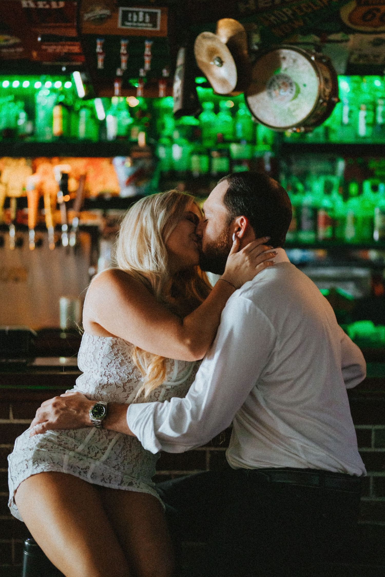 Couple sharing a romantic kiss at a bar with green-lit bottle display in the background.