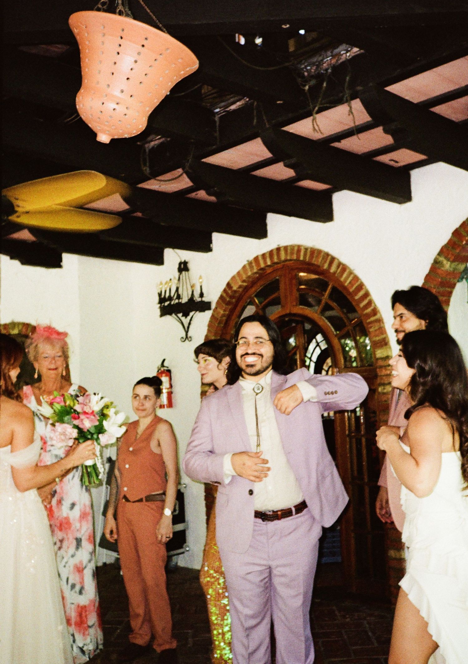 Groom in lavender suit laughing joyfully at wedding reception with guests in elegant venue.