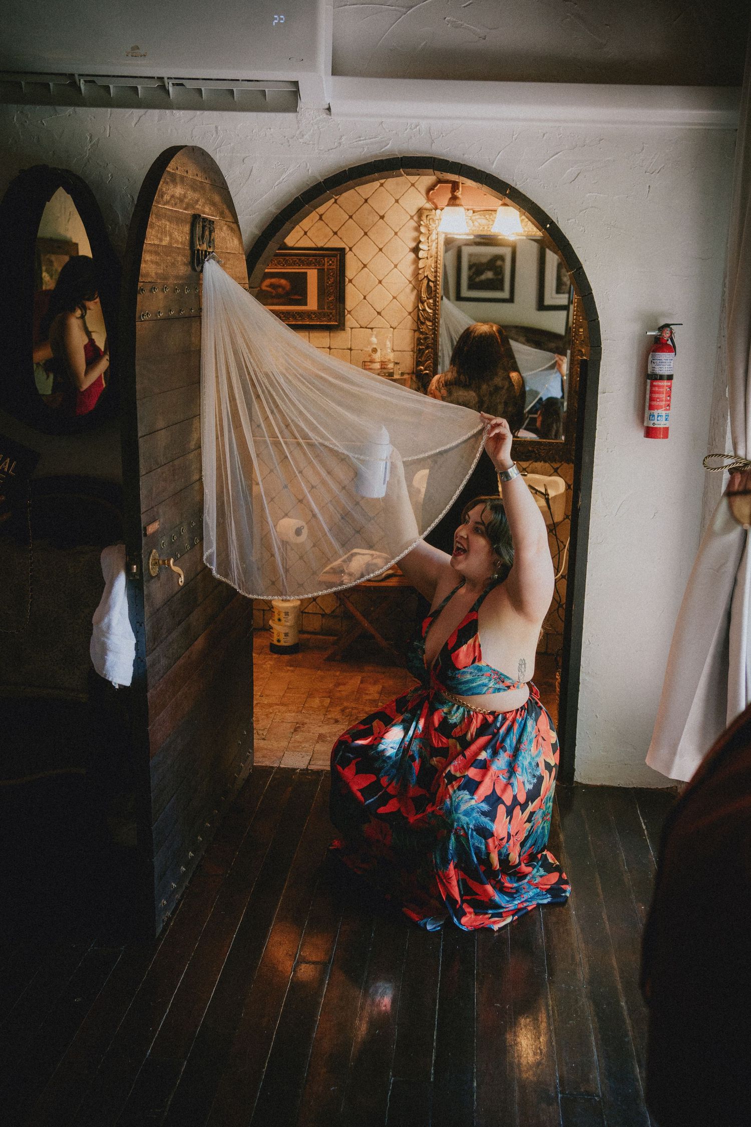 Woman in floral dress holding a wedding veil aloft in a rustic room with arched doorway and dark hardwood floors.