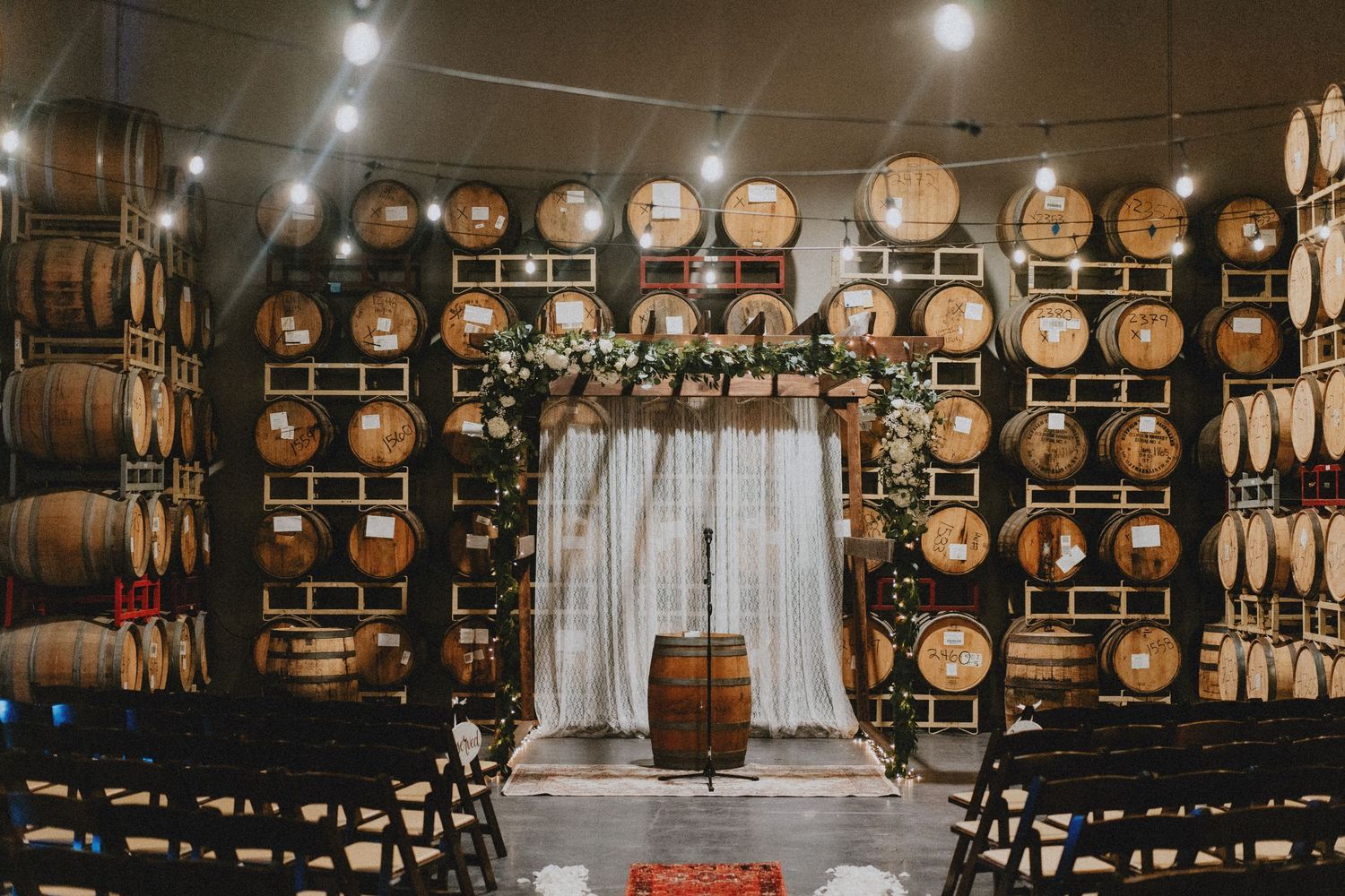 Rustic winery wedding ceremony setup with barrel walls, floral arch, string lights, and wooden chairs.