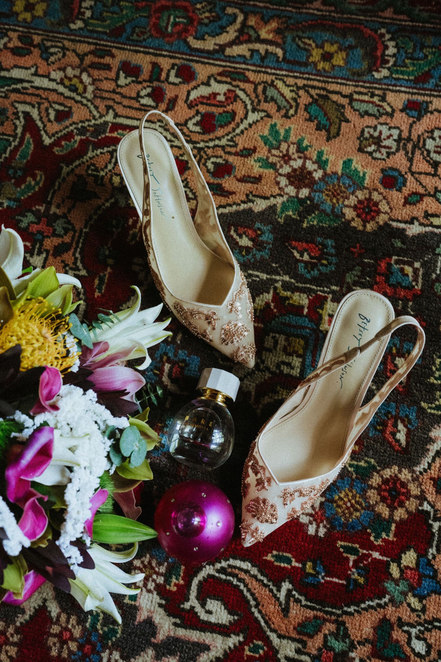 Floral slingback heels on a vintage rug beside a colorful bouquet and perfume bottles for a wedding flat lay.