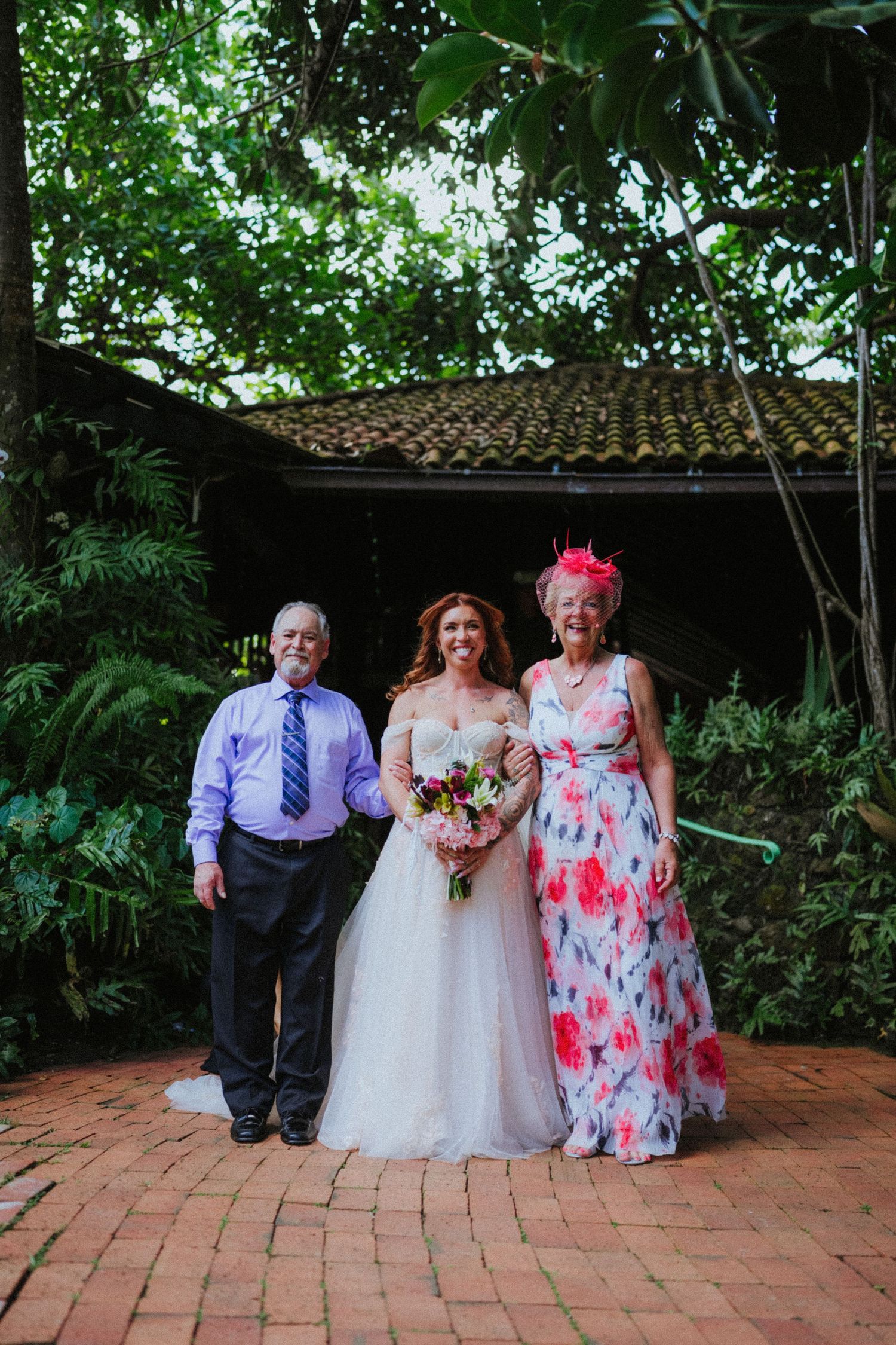 Bride in white gown poses with parents at tropical garden wedding venue, mother in floral dress.