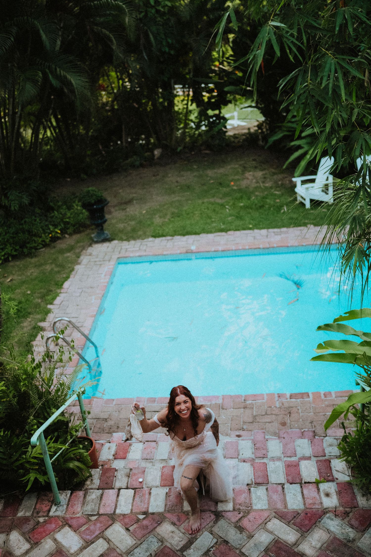 Woman in beige outfit sitting by a turquoise backyard pool surrounded by lush tropical greenery.