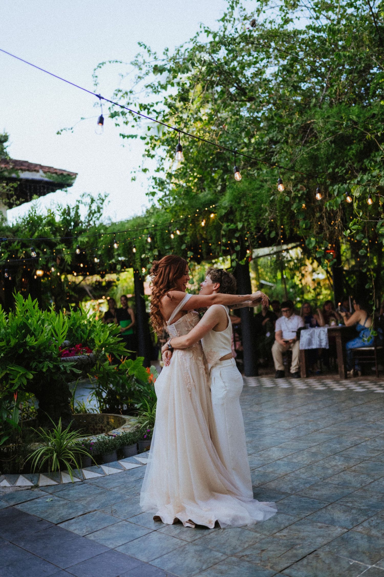 Two brides in white gowns share first dance at outdoor garden wedding with string lights and lush greenery.
