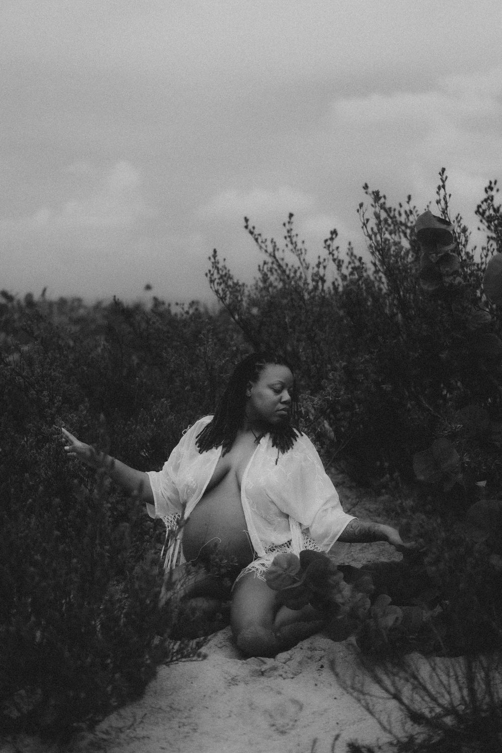 Artistic black and white portrait of a pregnant figure in white clothing kneeling on sand among bushes.