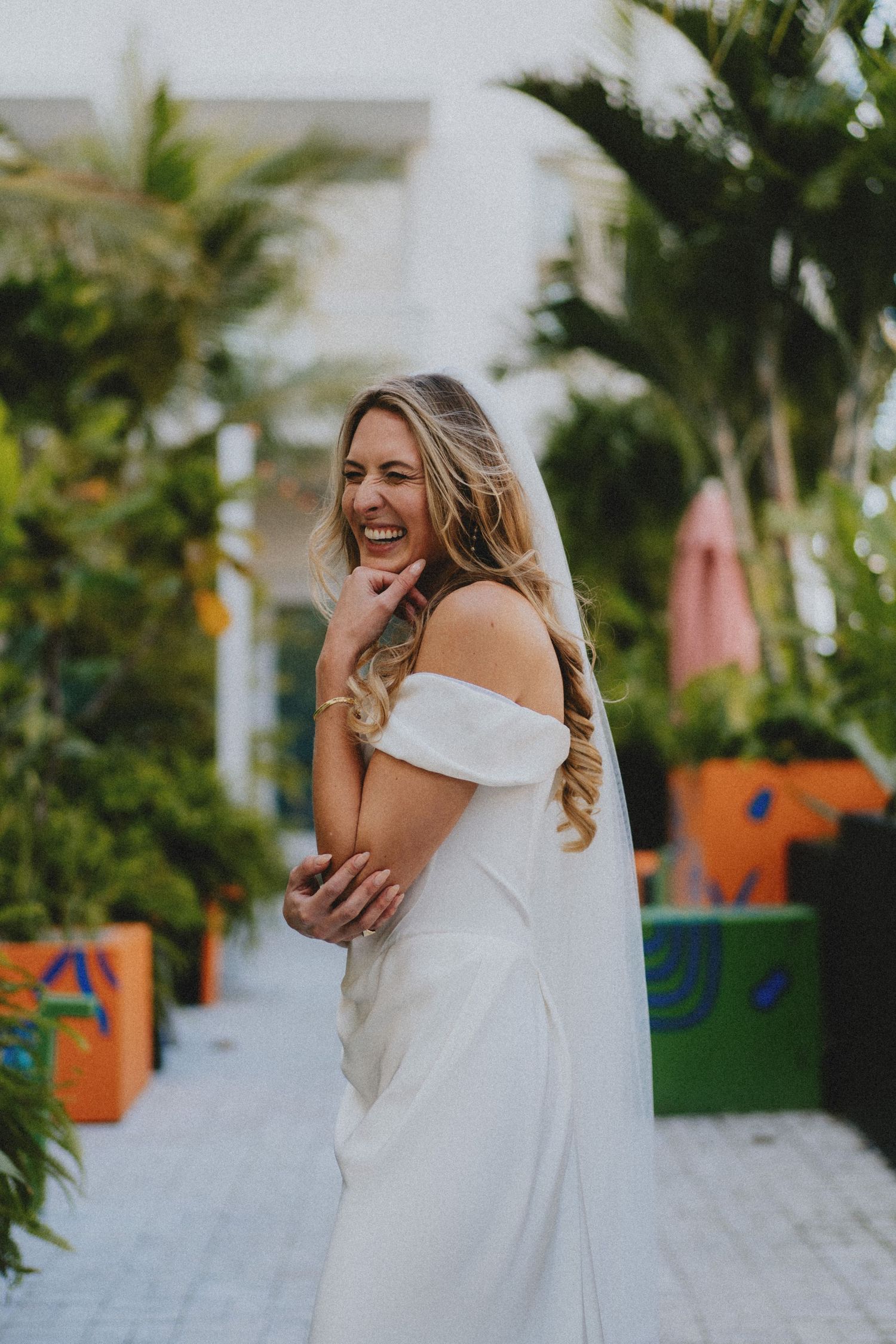 Smiling bride in off-shoulder white gown and veil laughing outdoors among tropical greenery.