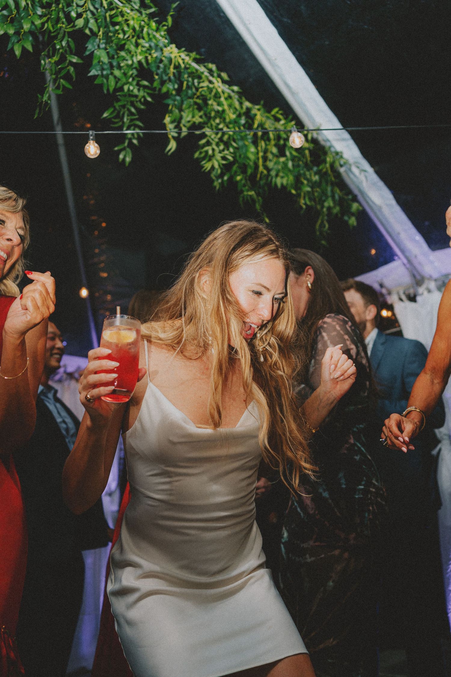 Bride in white slip dress dancing with drink in hand at outdoor wedding reception at night.