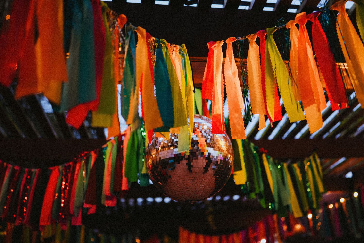 Colorful tassels and streamers hang from wooden beams above a glittering disco ball at a festive party.