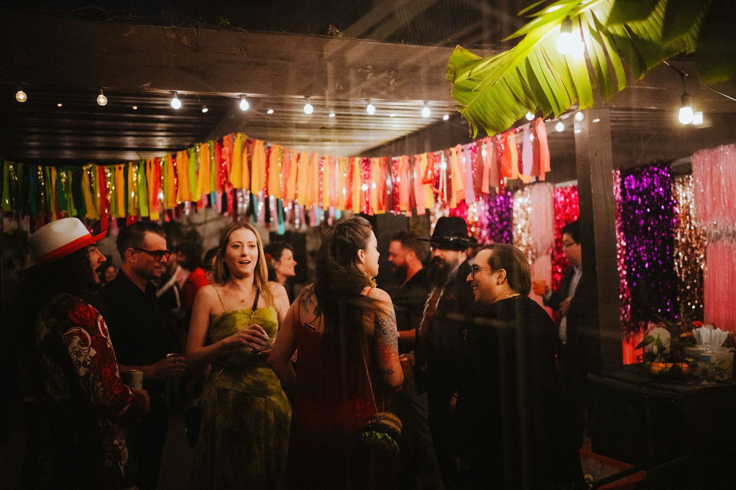 Guests mingle at a vibrant indoor party with colorful hanging decorations and warm string lights.