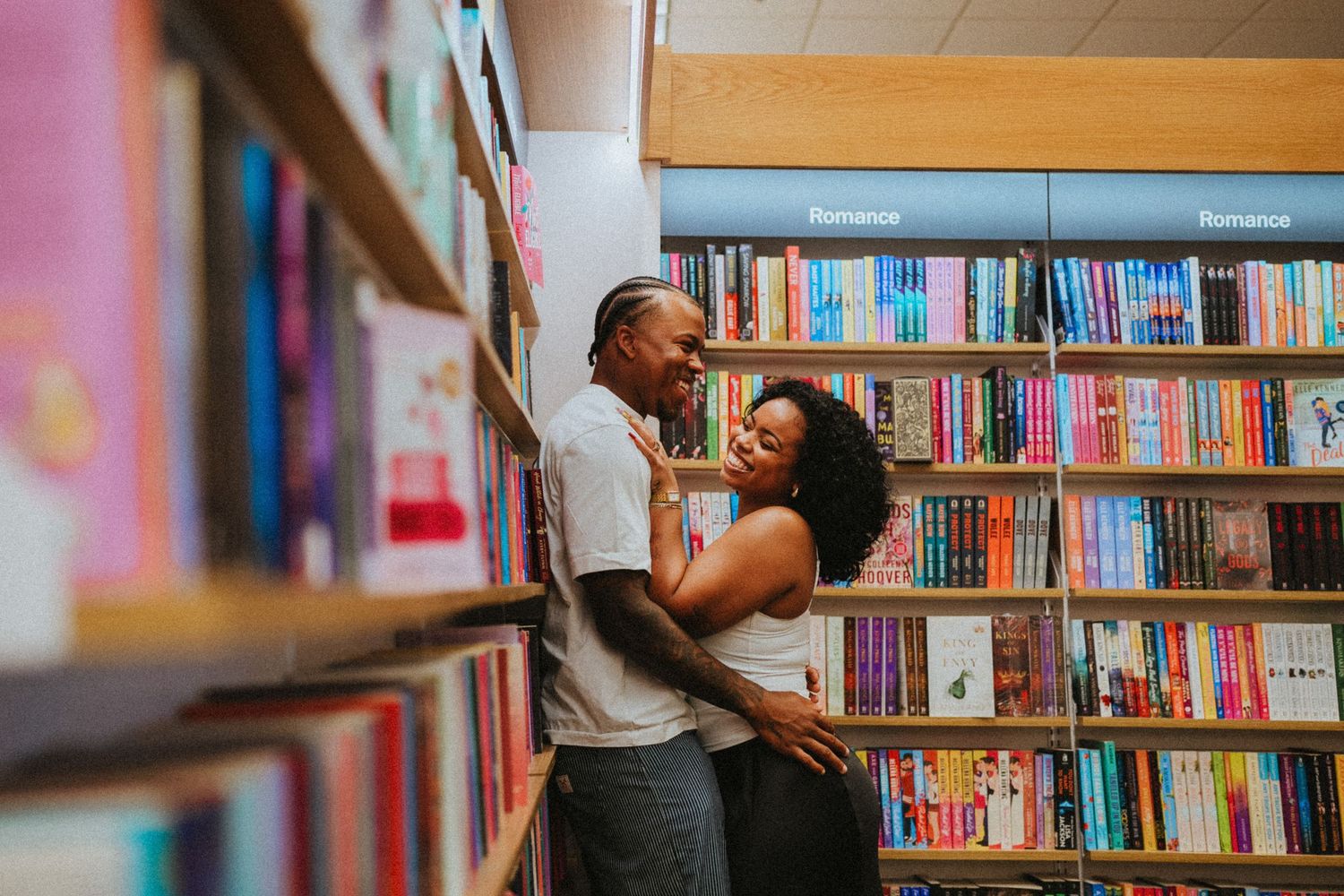 A couple shares a romantic embrace between bookshelves in a cozy bookstore, surrounded by colorful books.