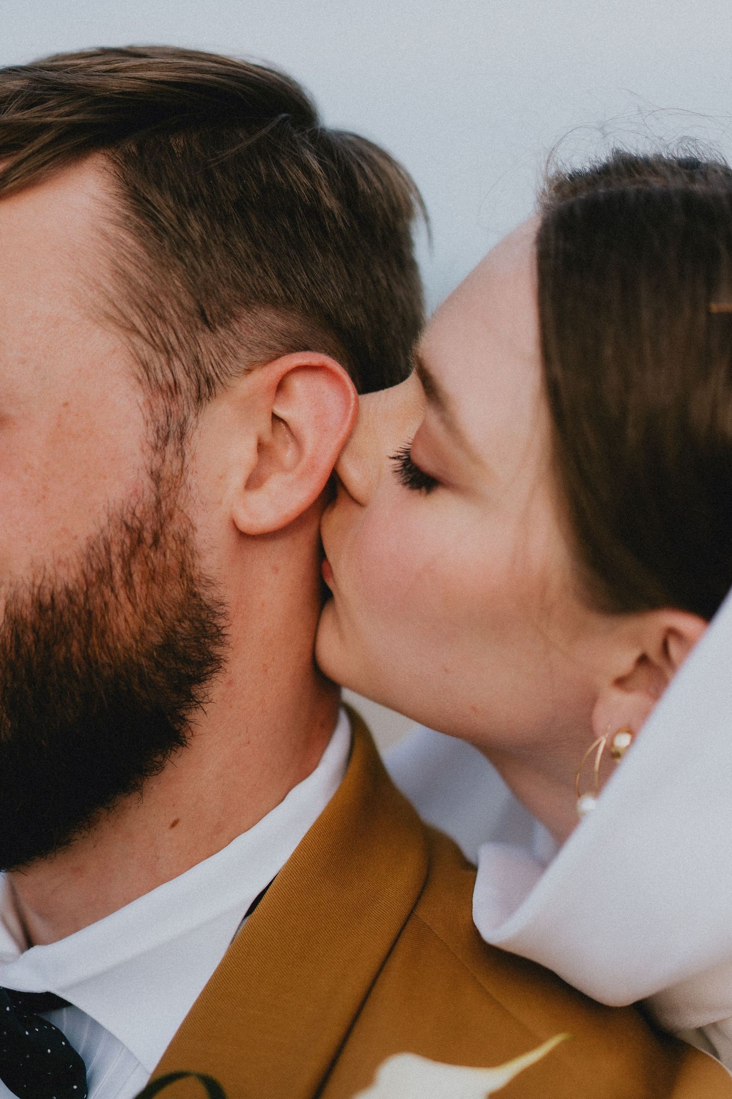 Close-up of bride kissing bearded groom in mustard suit on cheek.