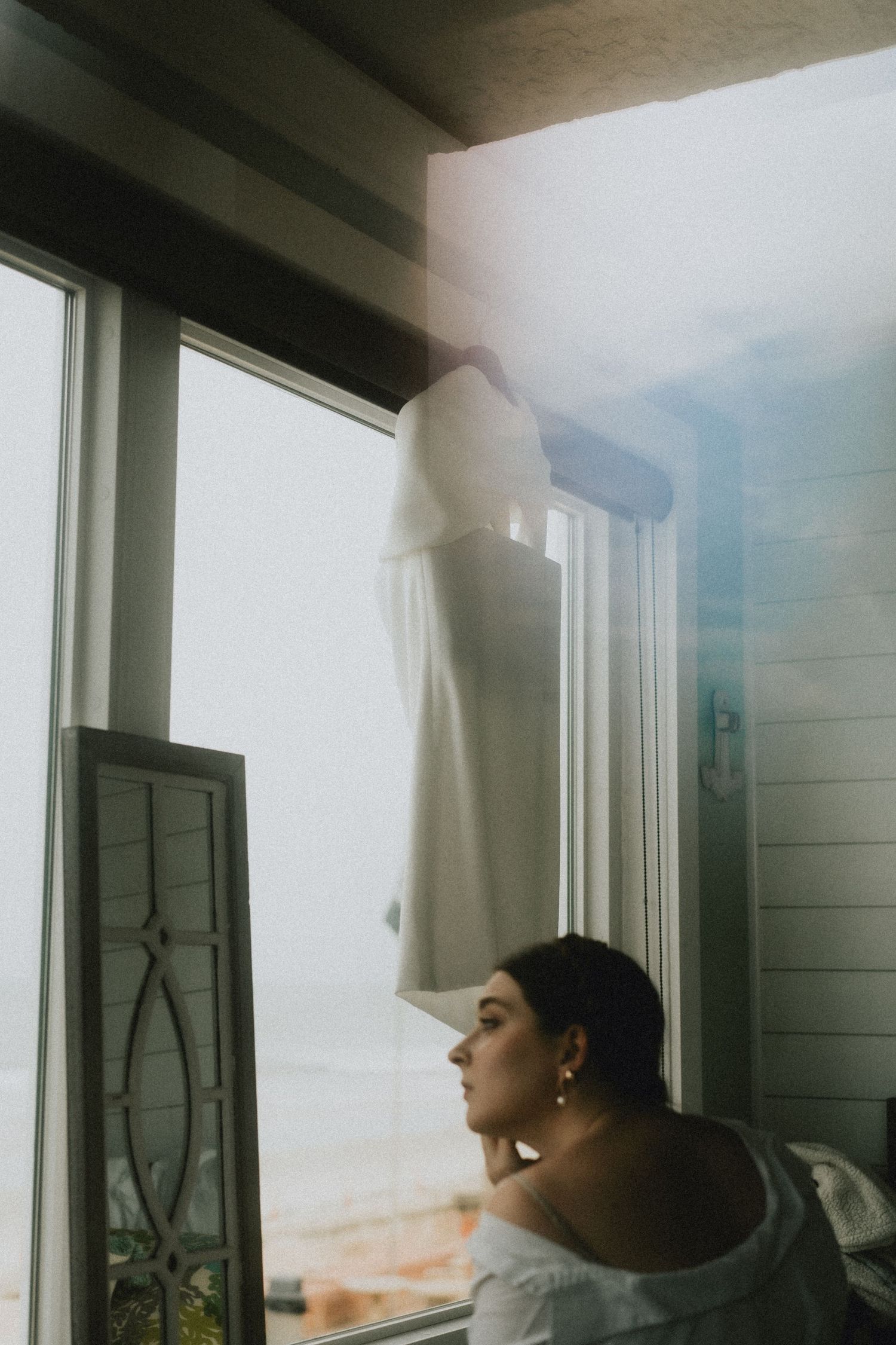 Bride gazing out window with wedding dress hanging nearby in soft natural light.