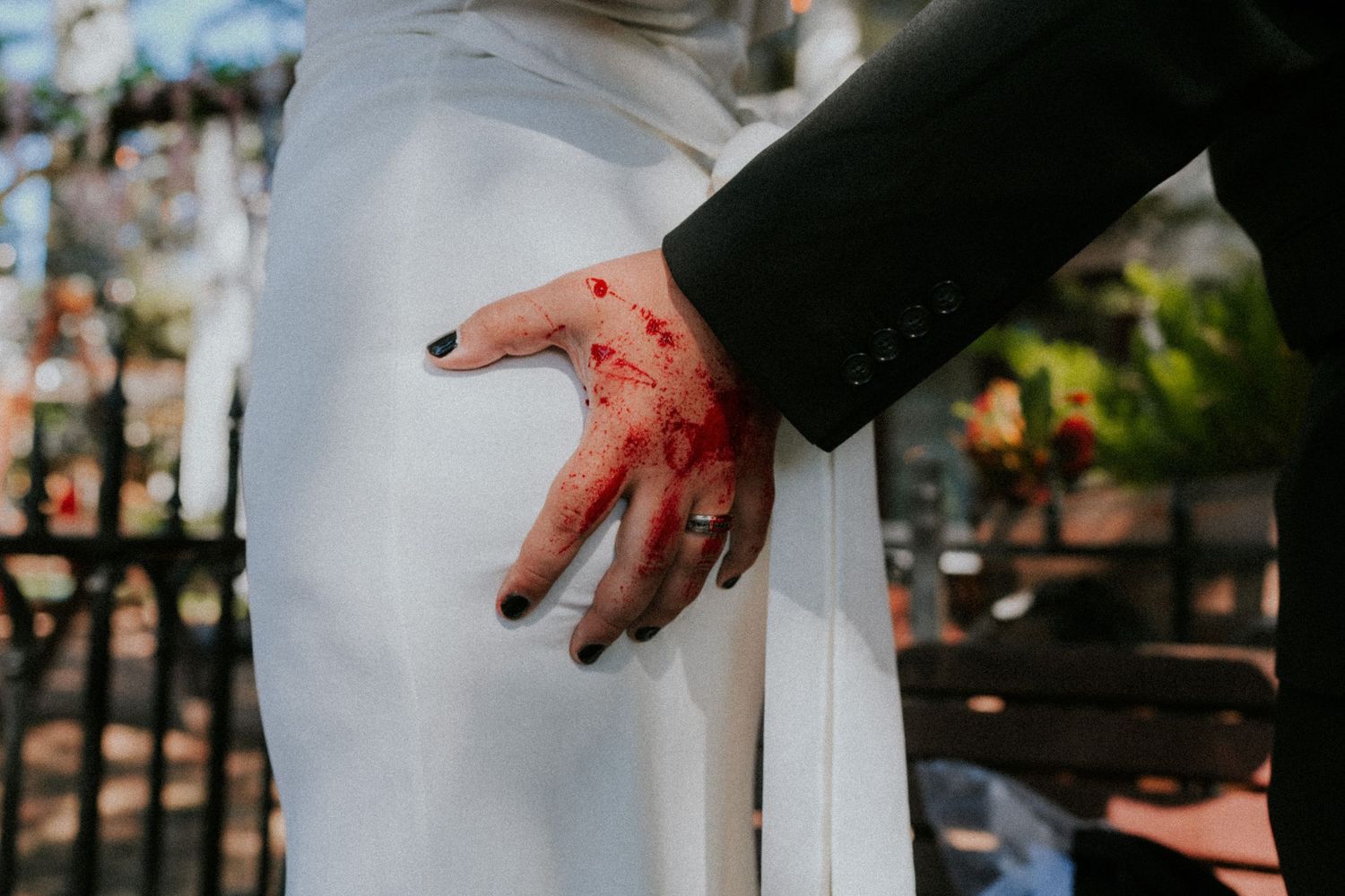 Close-up of bride and groom hands touching, with red floral detail on hand, wearing wedding attire and black nail polish.