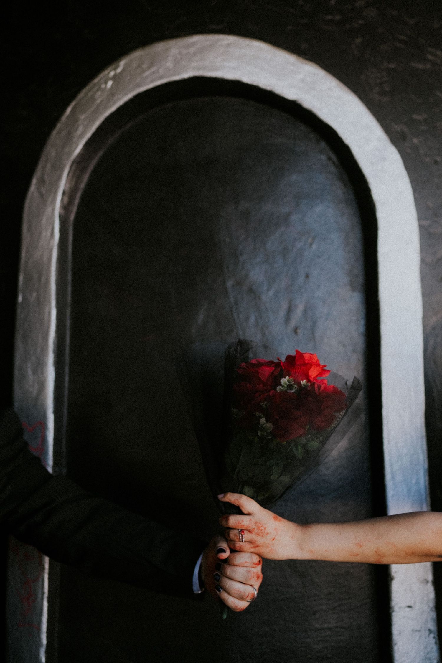 Hands holding a bouquet of red roses in front of a dark arched stone wall niche.