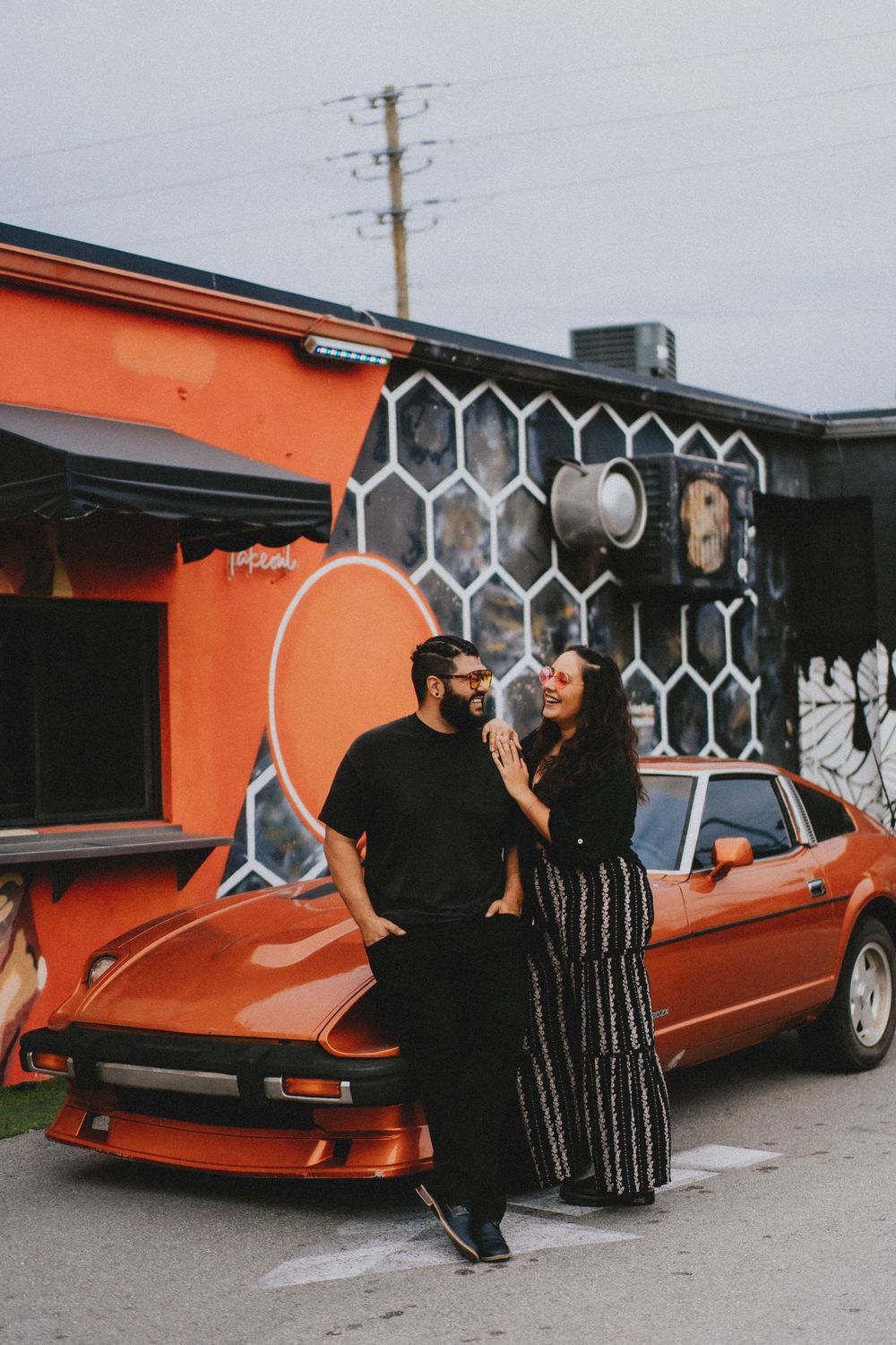Couple posing in front of orange wall with vintage car and geometric artwork.