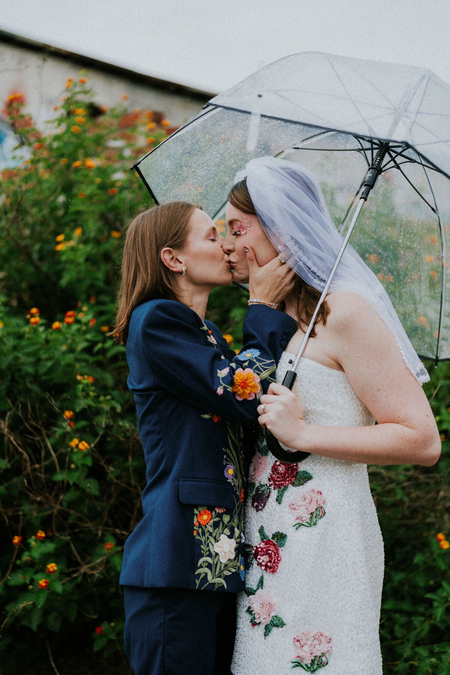 Two brides share a kiss under a clear umbrella, one in a floral suit and one in an embroidered wedding dress with veil.