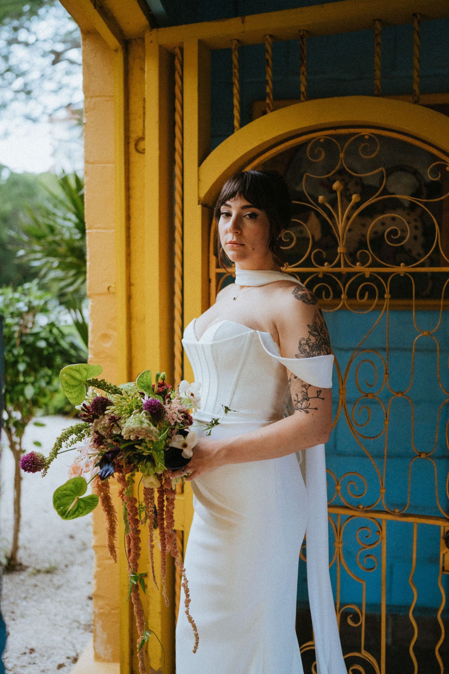 Tattooed bride in off-shoulder white gown holds a lush bouquet beside a yellow building with blue iron door.