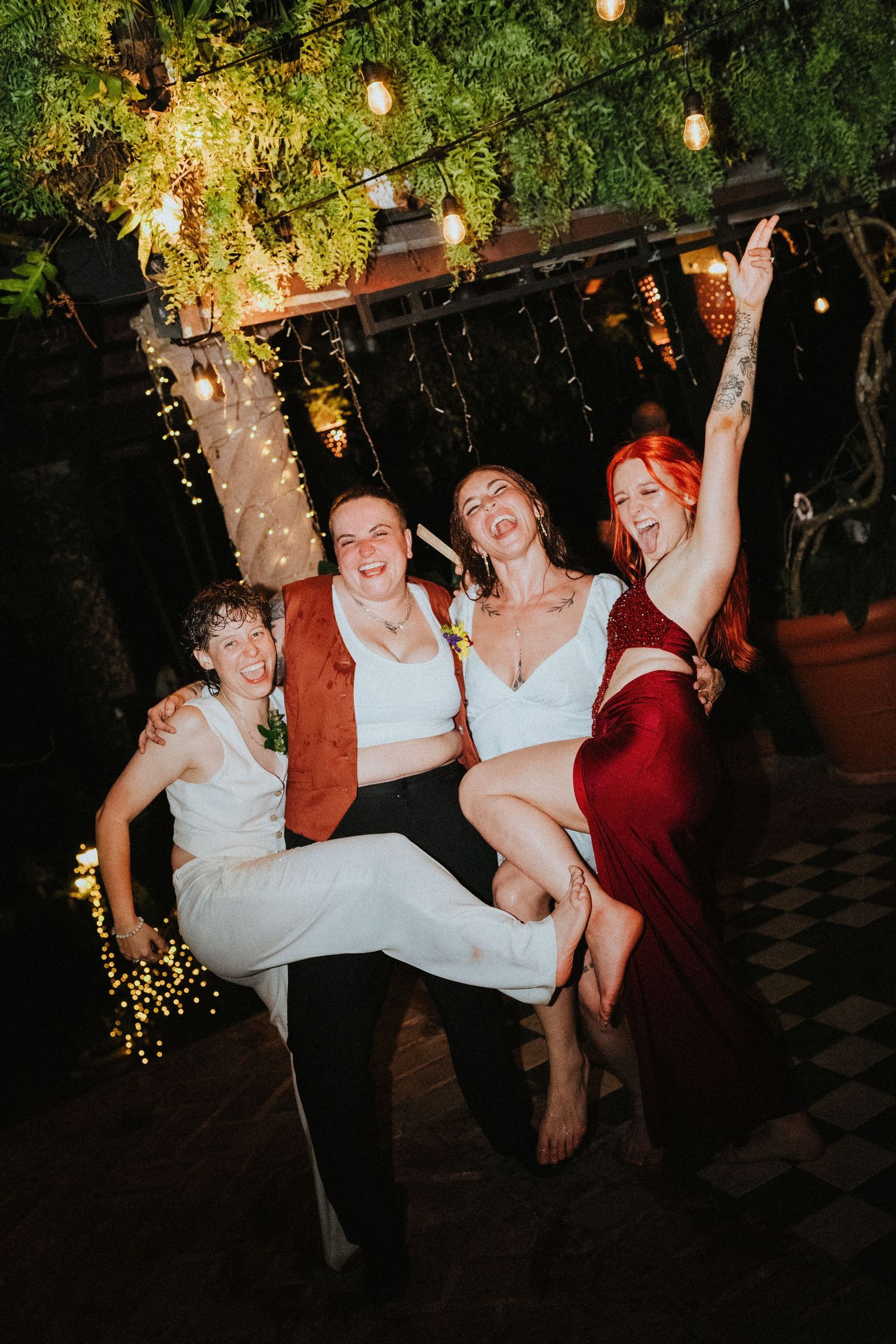 Four joyful women celebrate at a wedding reception, two lifting the bride in white, surrounded by string lights and lush greenery.