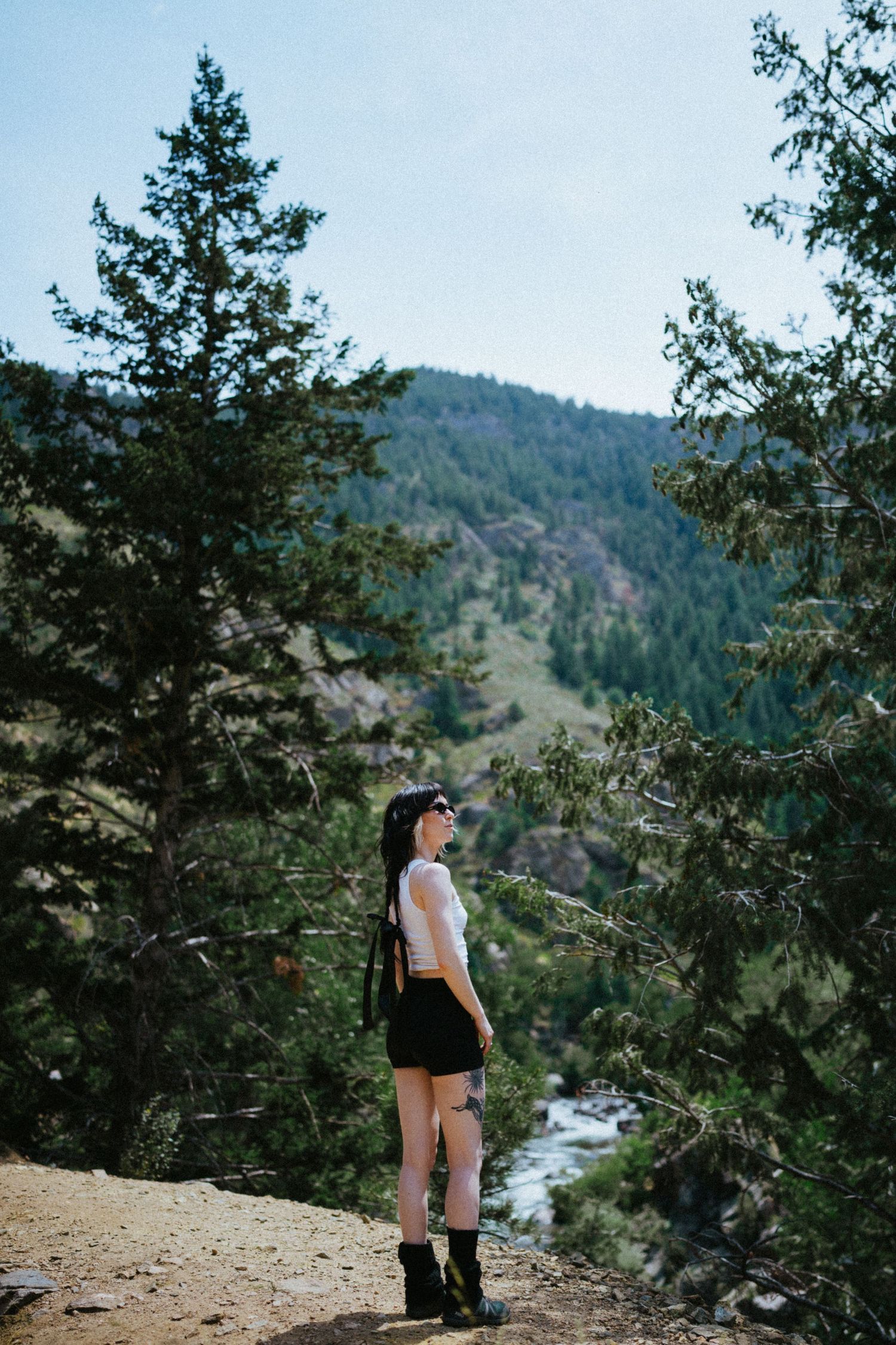 Young woman in white top and black shorts standing on a mountain trail with pine trees and river.