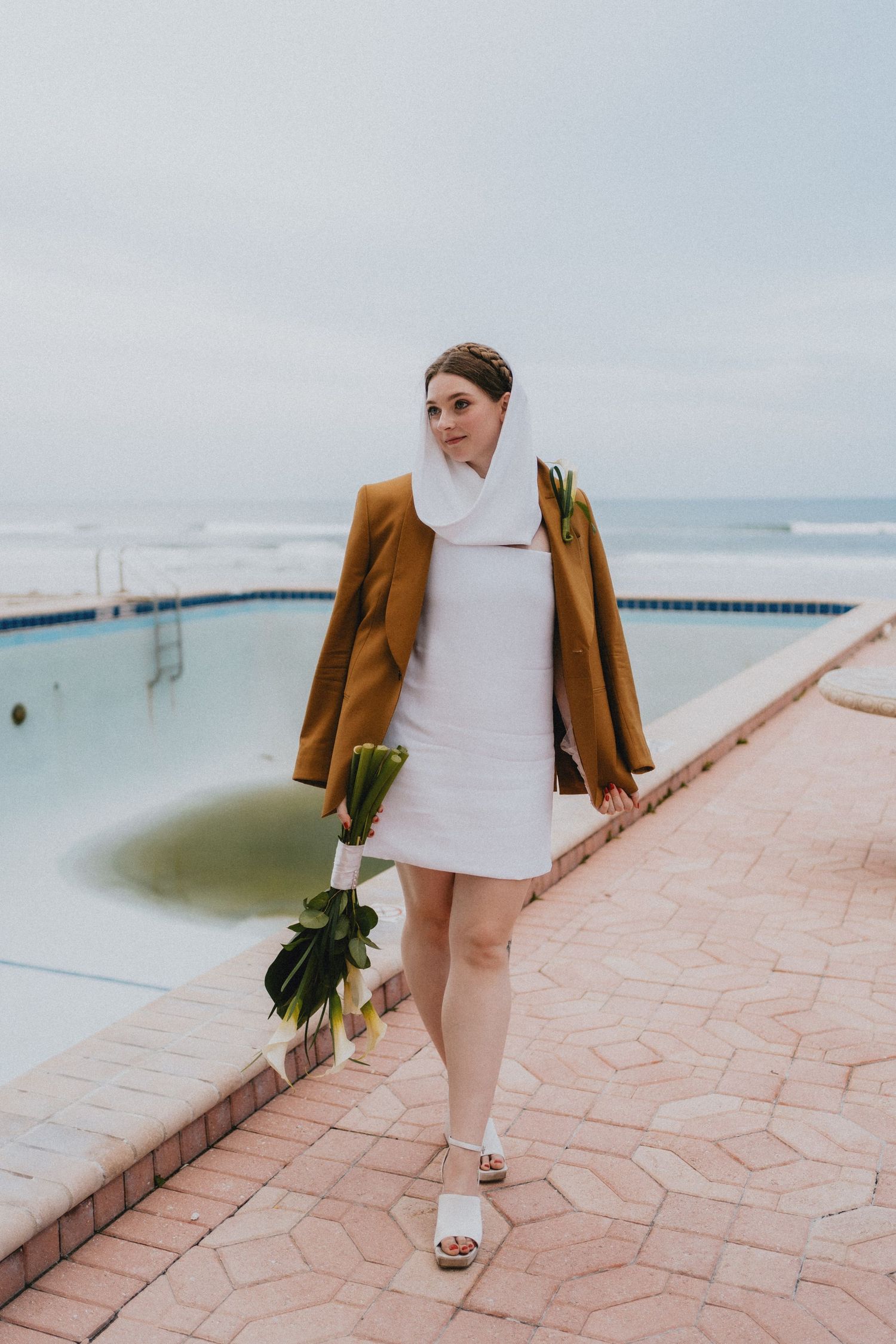 Bride in white mini dress and mustard blazer holding tropical bouquet by pool.