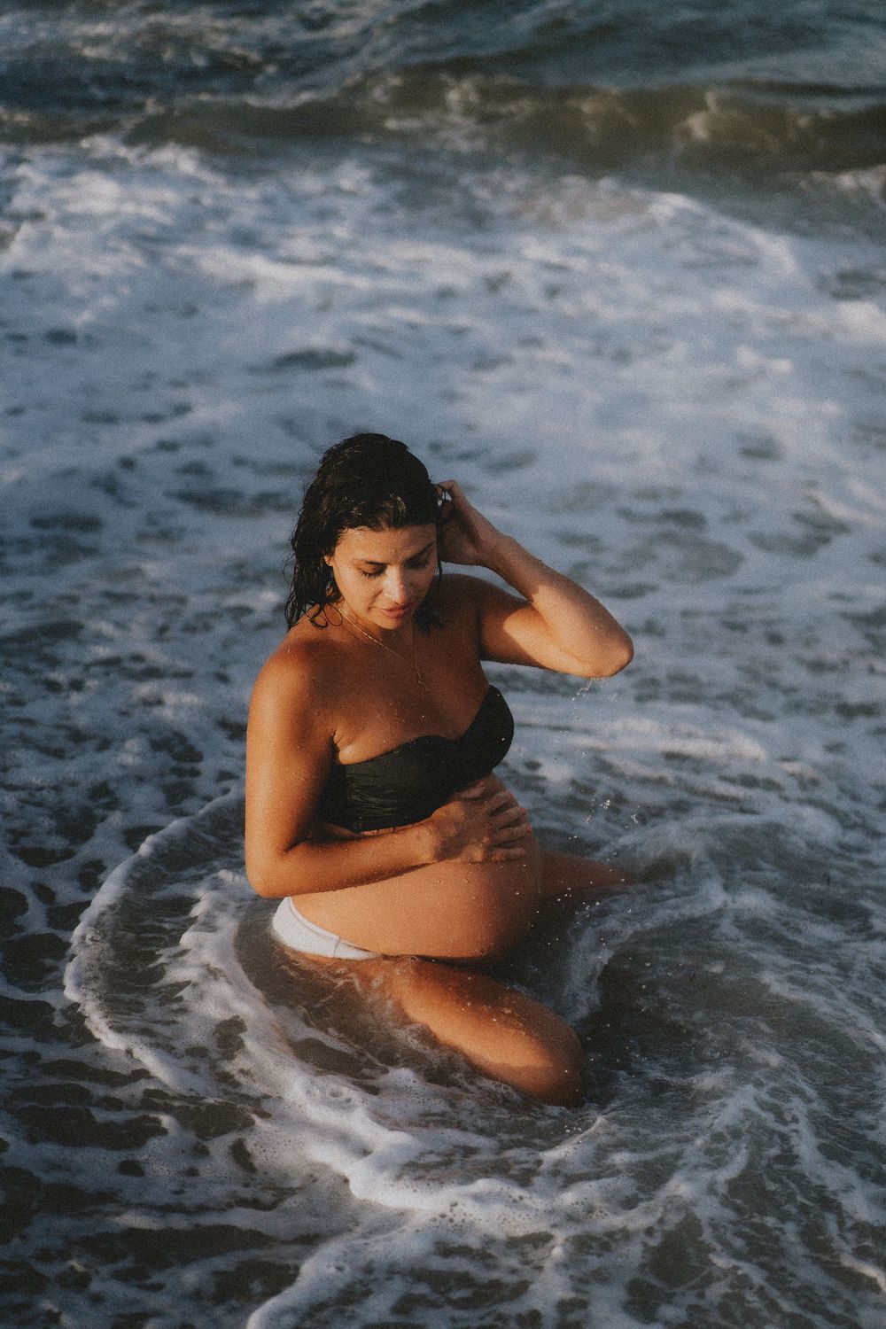 A figure in a black swimsuit sits in shallow ocean waves during golden hour lighting.