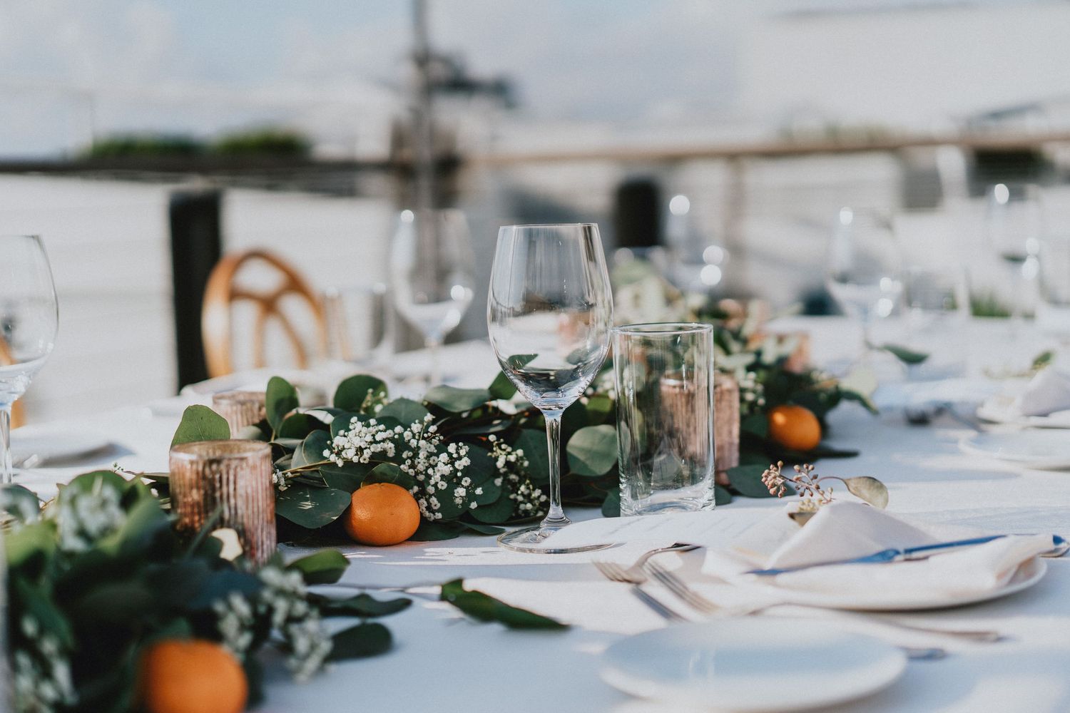 Elegant outdoor wedding table with greenery garland, oranges, candles, and crystal glasses on white linen.