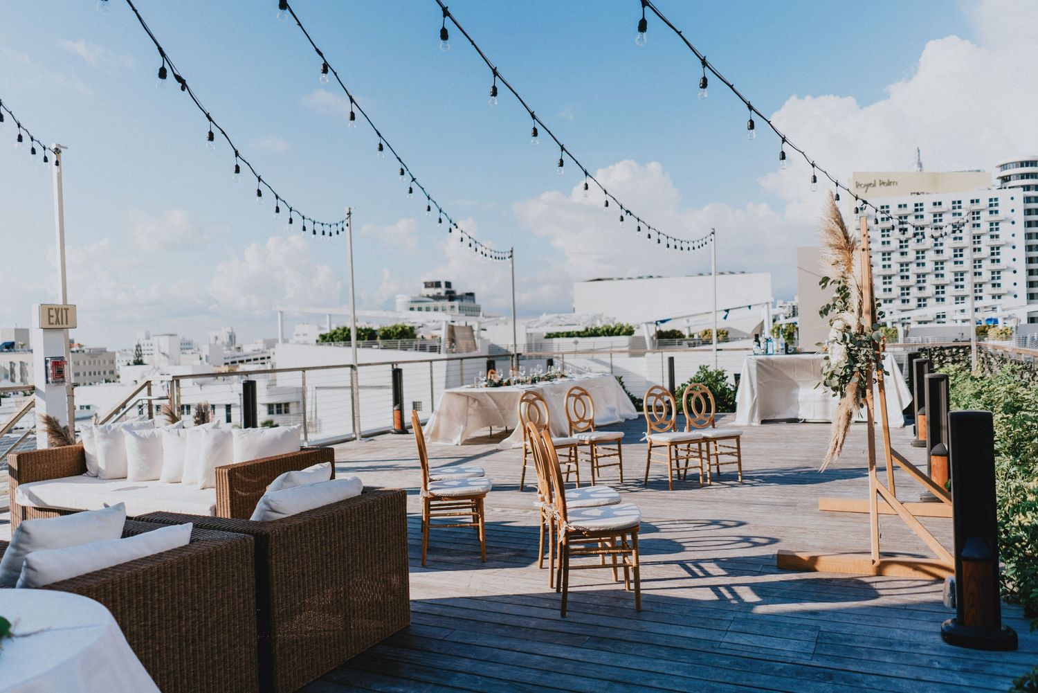 Rooftop venue with string lights, wicker furniture, and wooden deck overlooking city skyline on a sunny day.