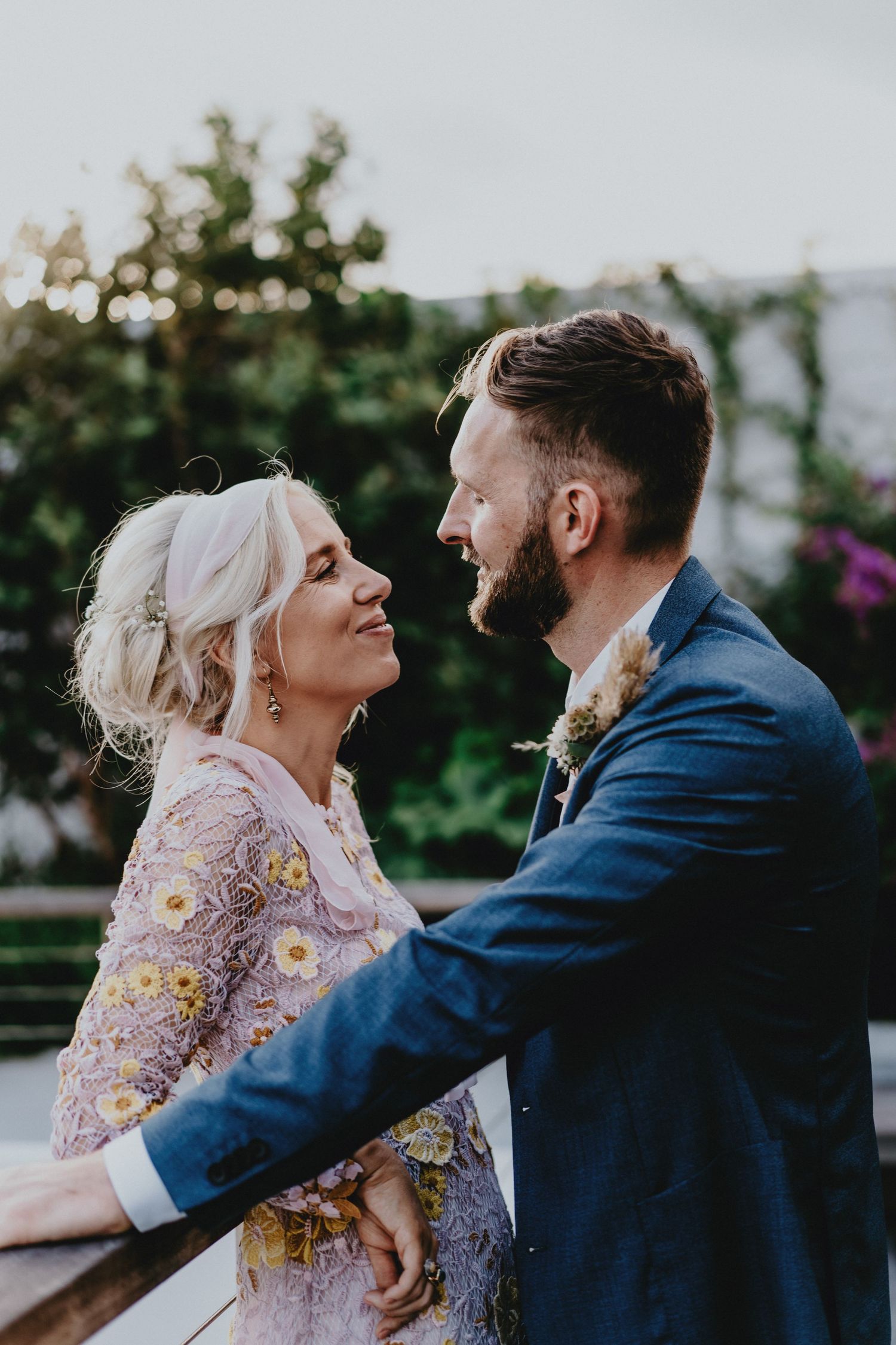 A bride in a floral dress gazes lovingly at a bearded groom in a blue suit on a balcony surrounded by greenery.