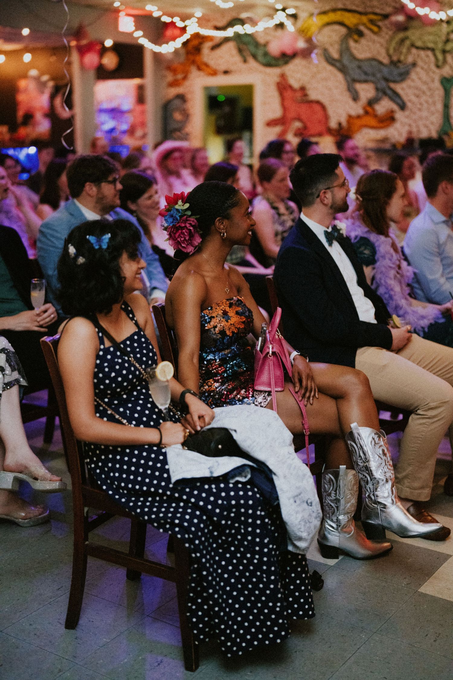 Guests in colorful attire sit watching a wedding ceremony in a vibrant, eclectic venue decorated with festive lights.