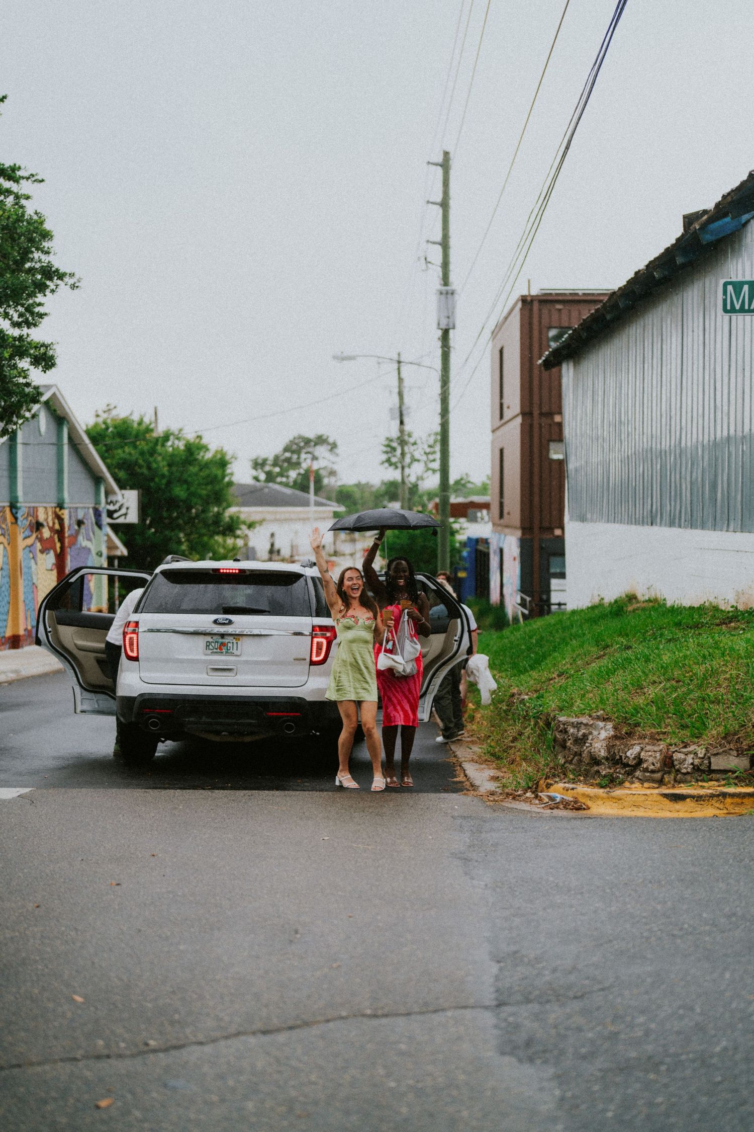 Two women in colorful dresses stand beside a white Ford SUV on a rainy urban street.