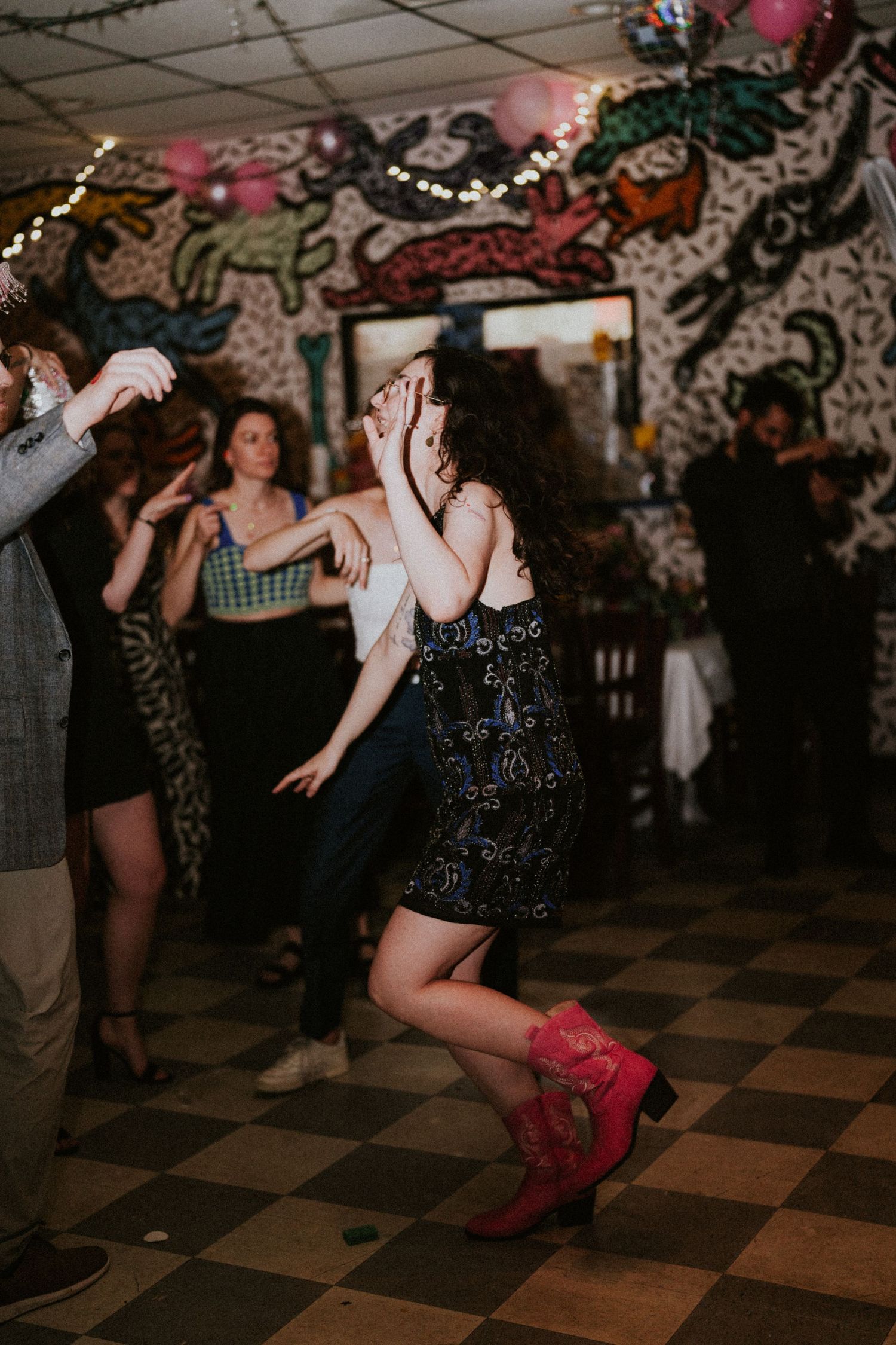 Woman in sequin dress and red cowboy boots dancing energetically on a checkered dance floor.