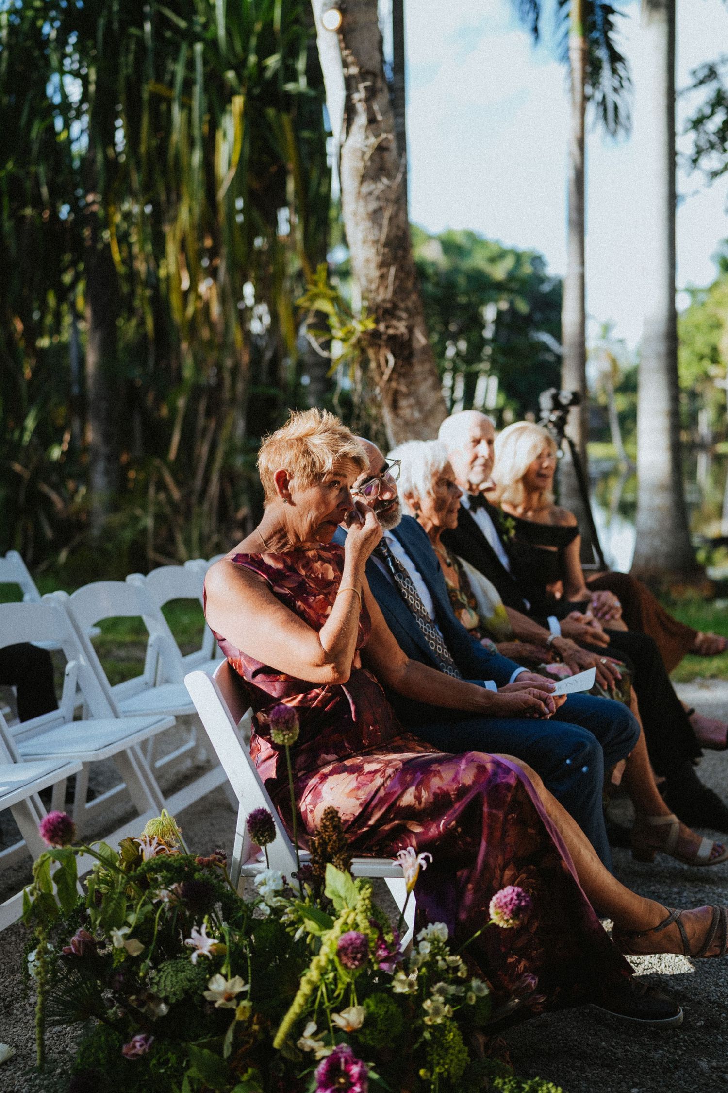 Emotional wedding guests seated in white chairs outdoors, surrounded by tropical palms and floral arrangements.