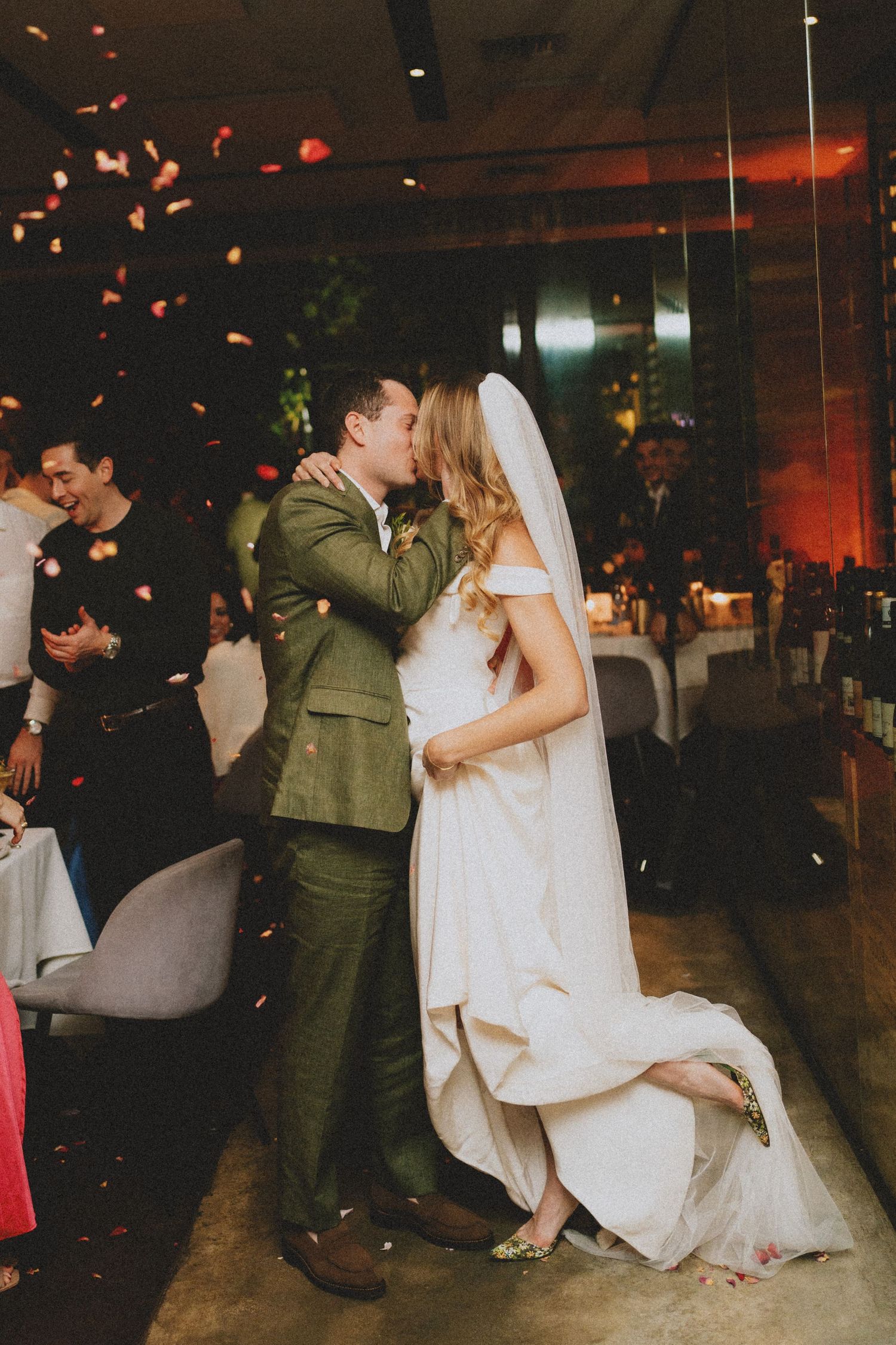 Bride and groom sharing their first kiss as wedding guests toss rose petals at the reception.