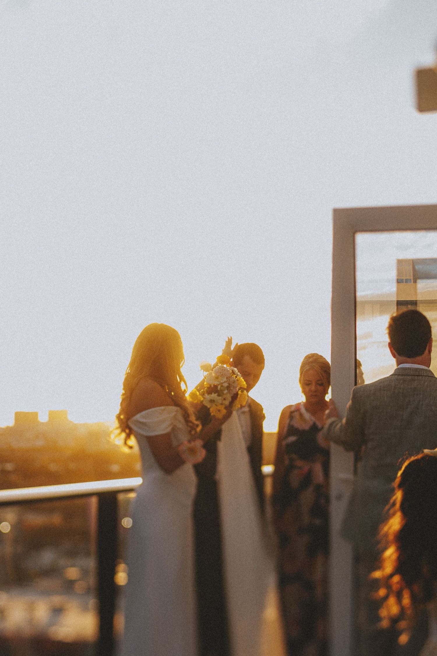 Bride holding bouquet silhouetted against golden sunset sky during outdoor rooftop wedding ceremony.