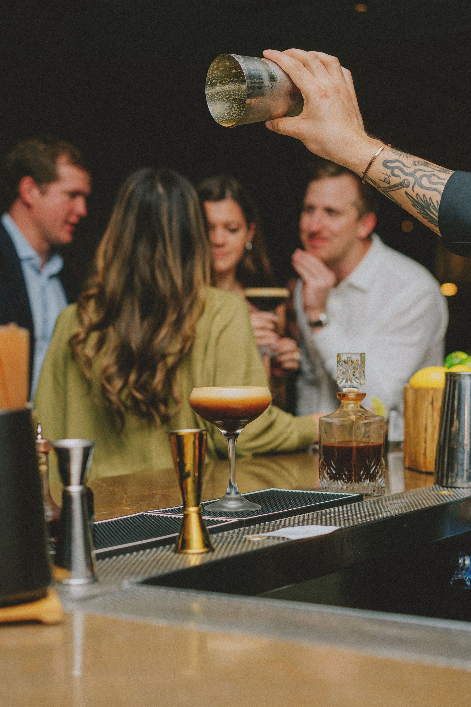 Tattooed bartender shaking a cocktail behind a bar with espresso martini and guests in the background.