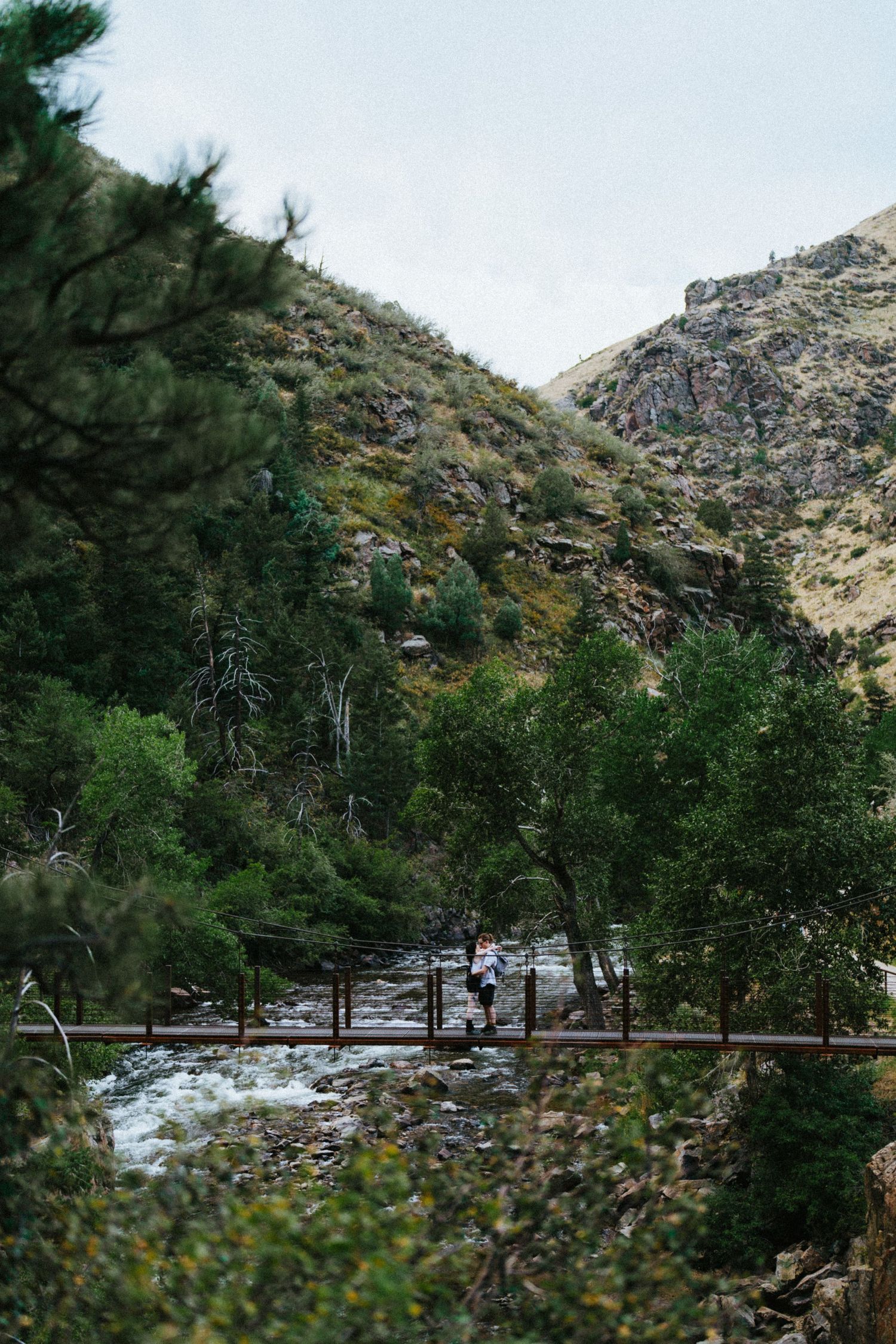 Couple embracing on a wooden footbridge over a rushing mountain river surrounded by lush green trees.