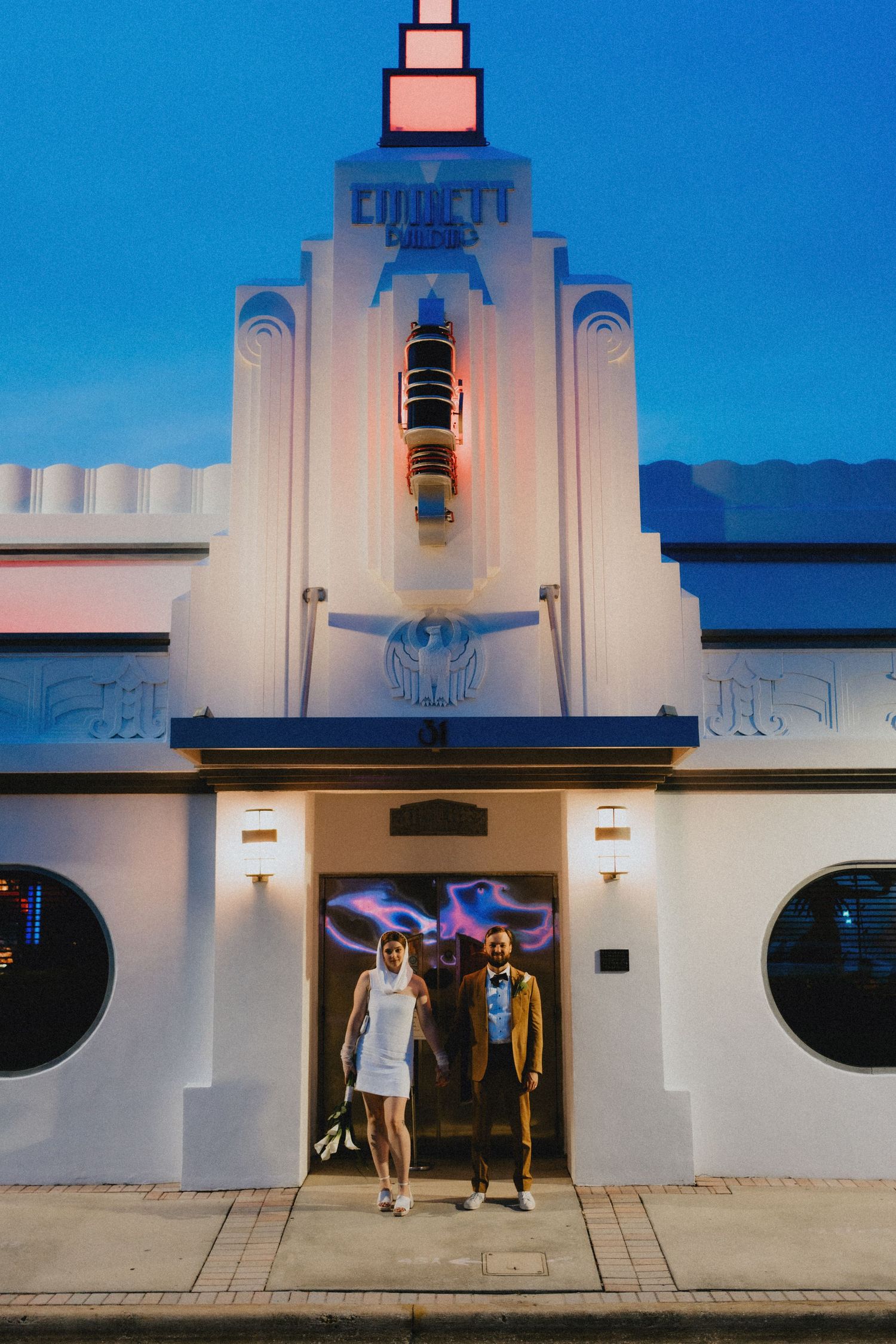 Bride and groom exit art deco venue at dusk, bride in white mini dress, groom in tan suit.