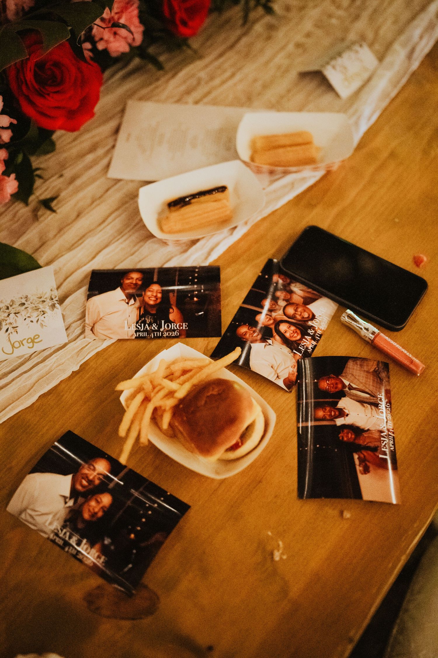 Wedding reception table with photos, smartphone, lip gloss, fries, and roses decorating a wooden surface.