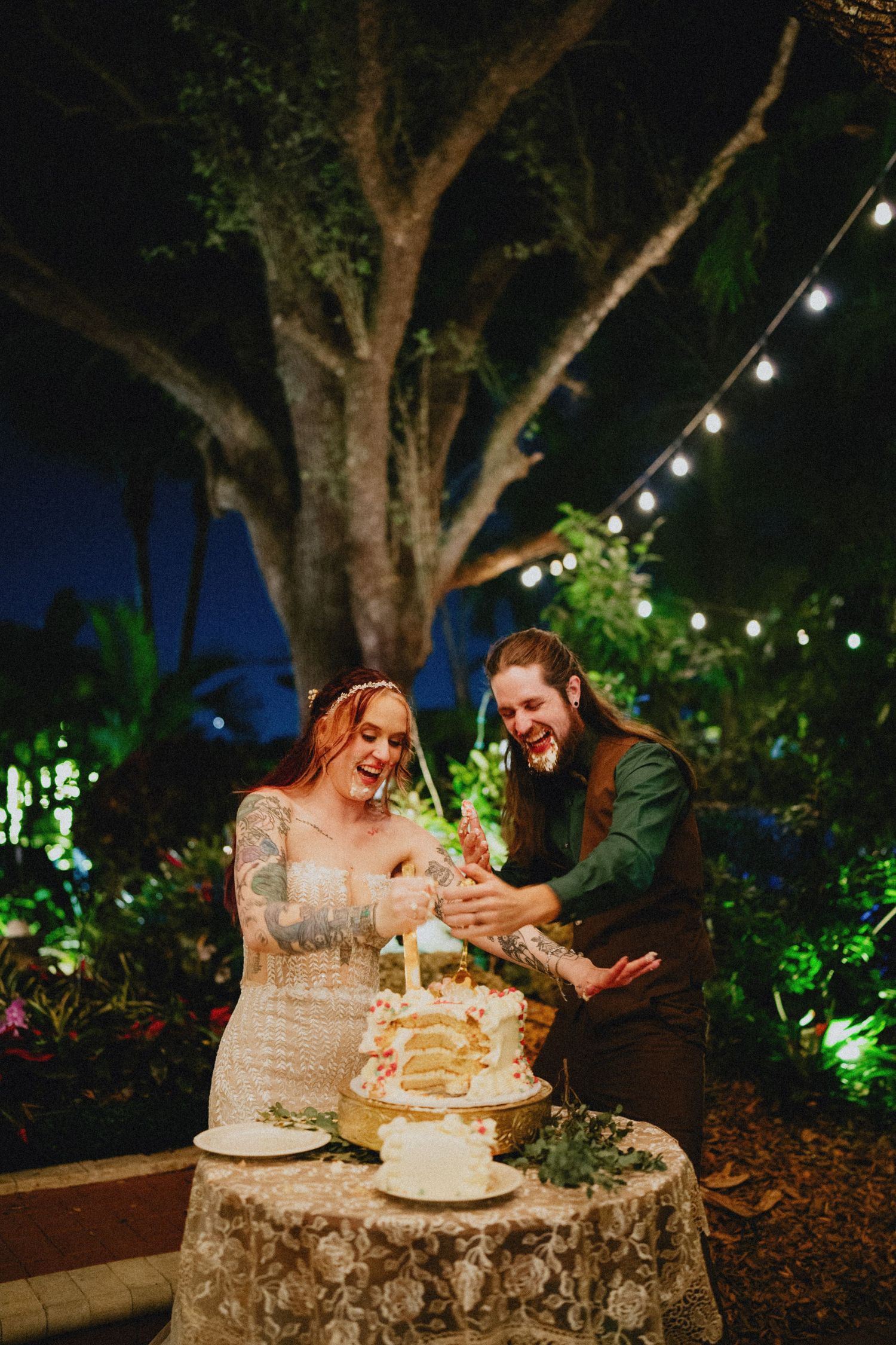 Bride and groom laughing while cutting wedding cake outdoors at night under string lights and lush tropical greenery.