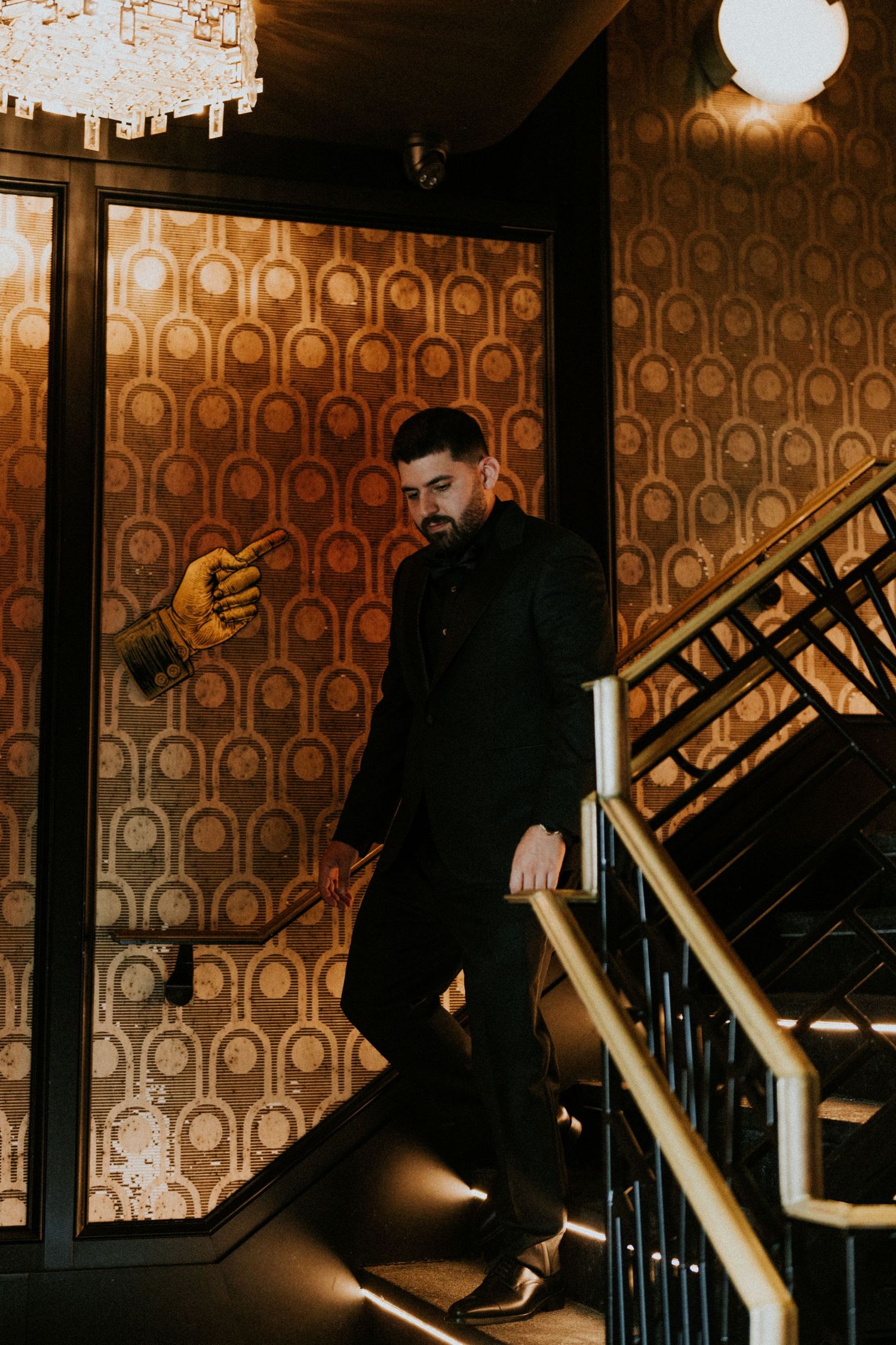 Stylish man in black suit descending ornate staircase in vintage gold patterned interior.