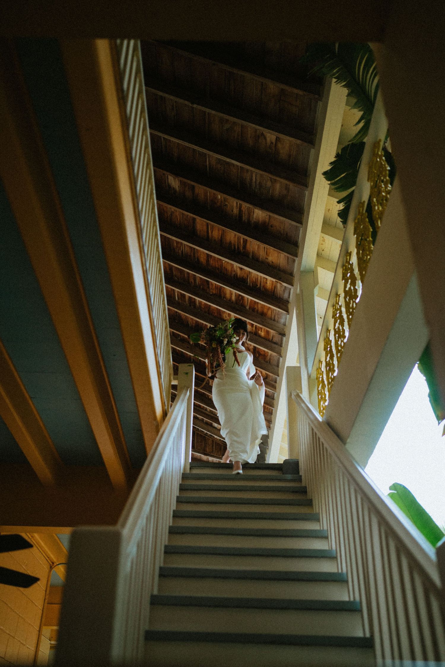 Bride in white gown descends a staircase with wooden ceiling in an elegant historic venue.