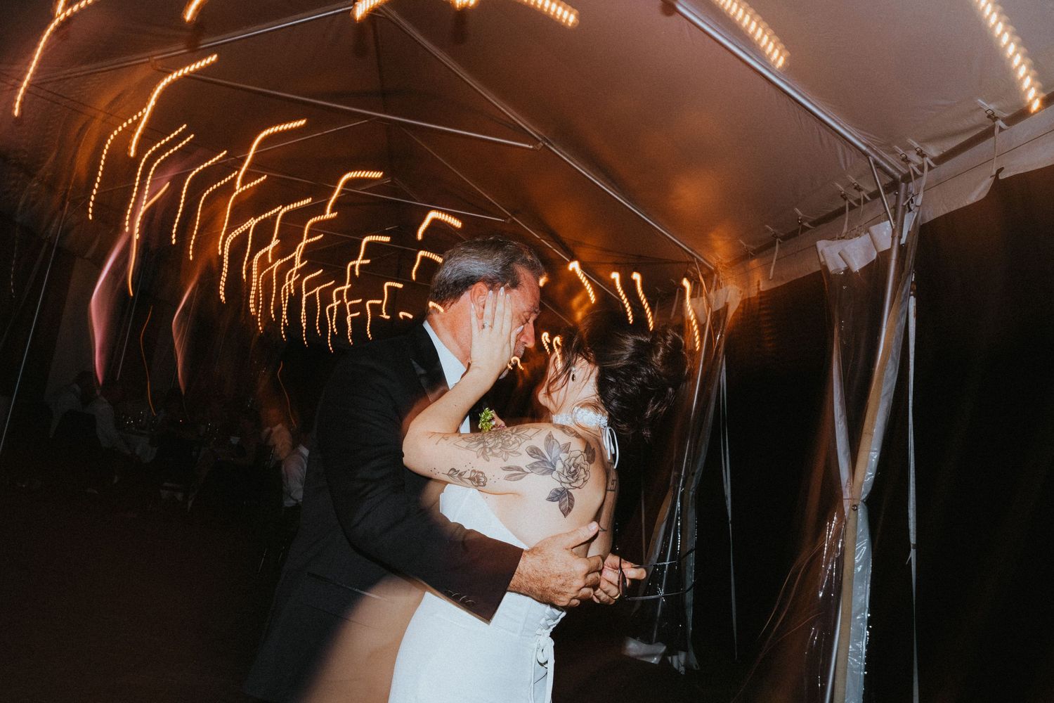 Bride and groom share a passionate kiss on the dance floor under glowing string lights at their wedding reception.