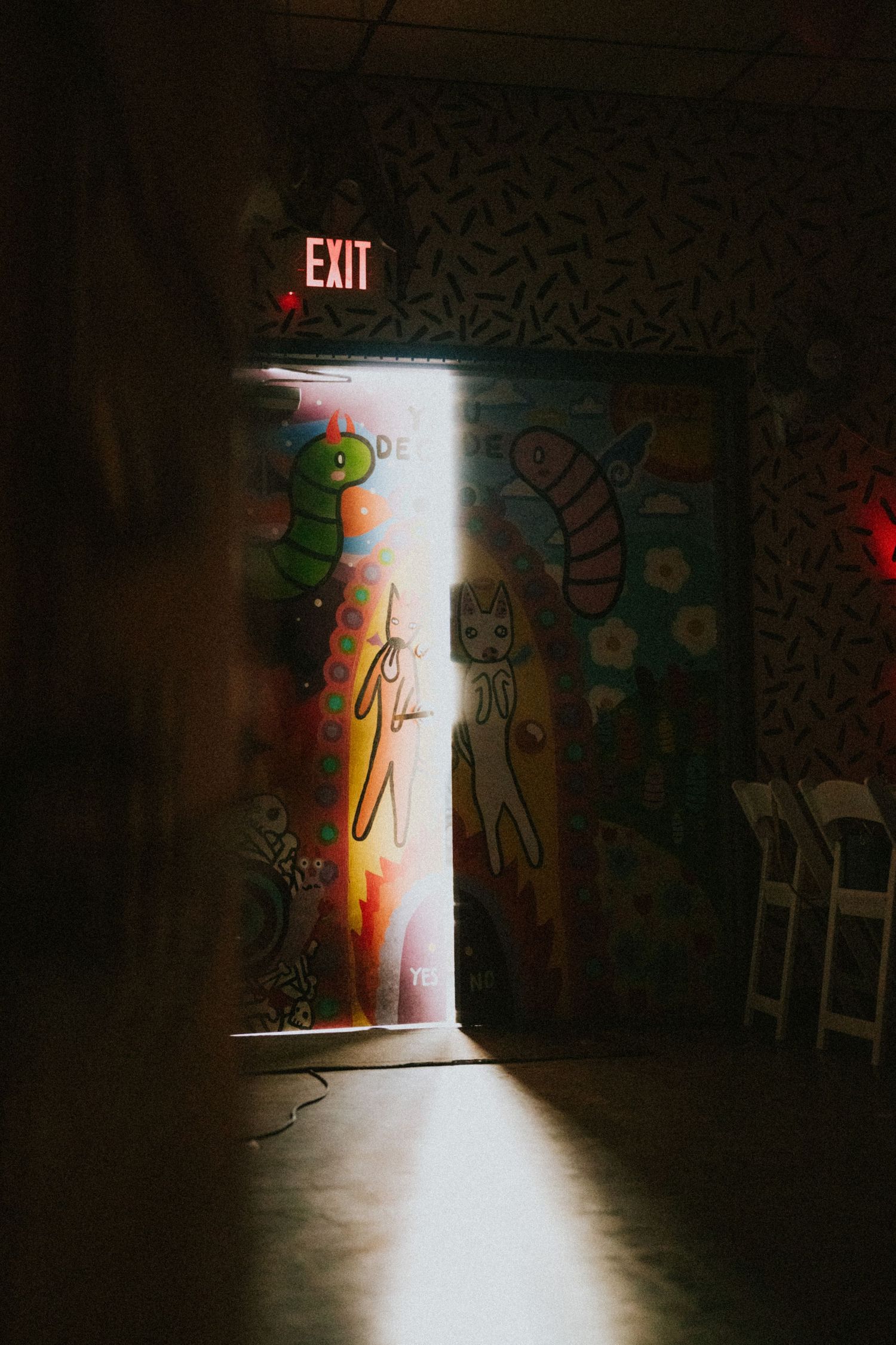 Colorful graffiti mural surrounds a glowing exit door in a dark room, featuring cartoon characters and vibrant artwork.