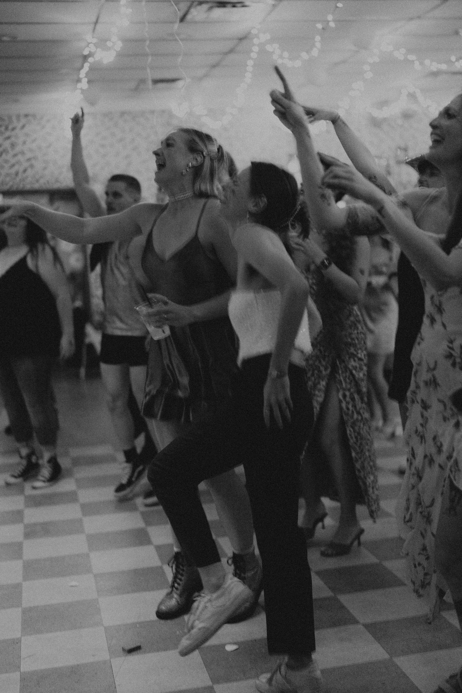 Black and white photo of guests dancing joyfully at a wedding reception on a checkered floor.